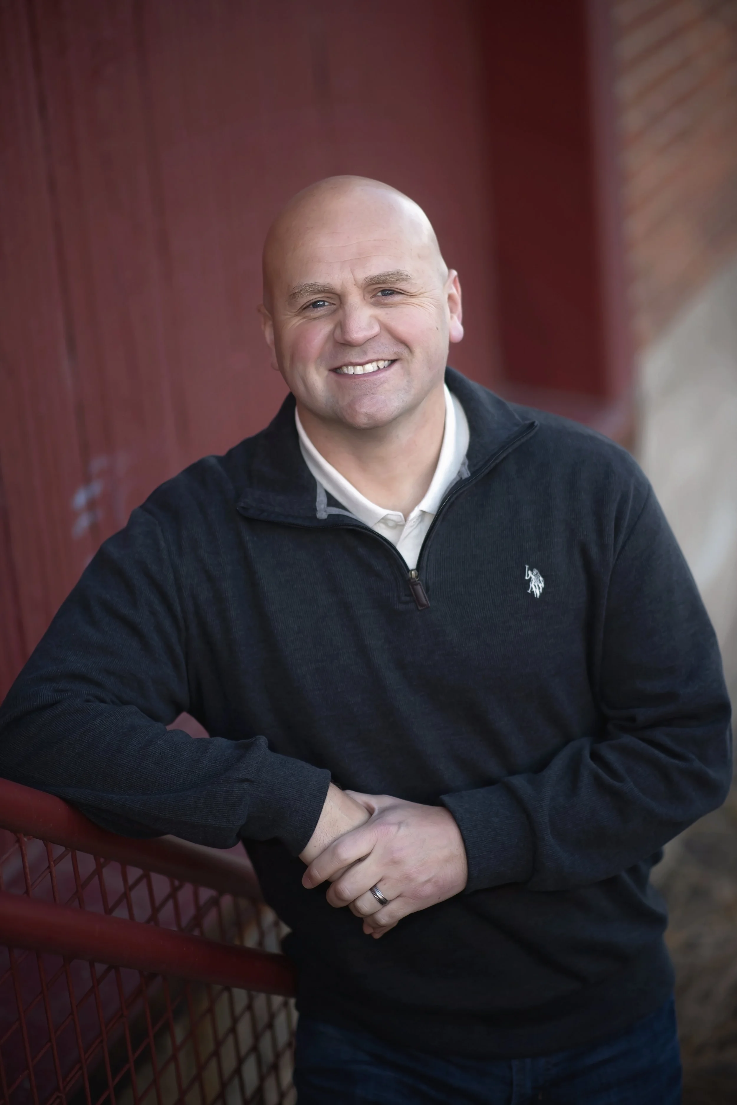 A smiling bald man in a black jacket leaning against a red metal fence outdoors.