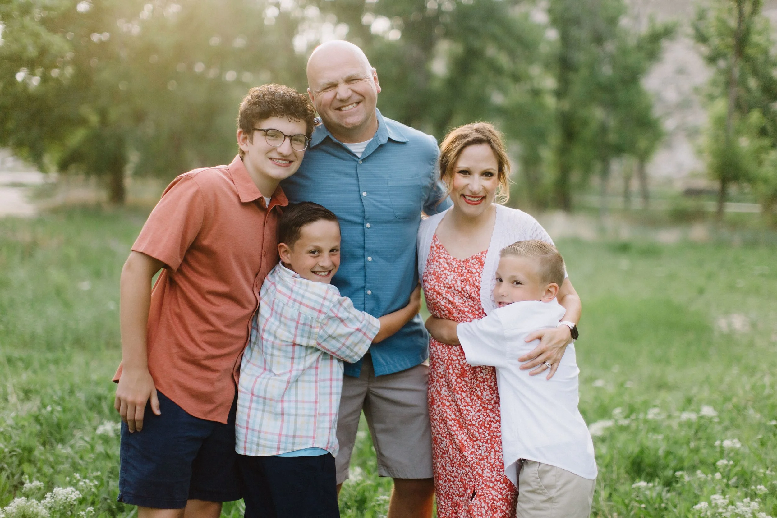 A family of six, including two adults and four children, standing outdoors on a grassy field with trees in the background, smiling and hugging each other.