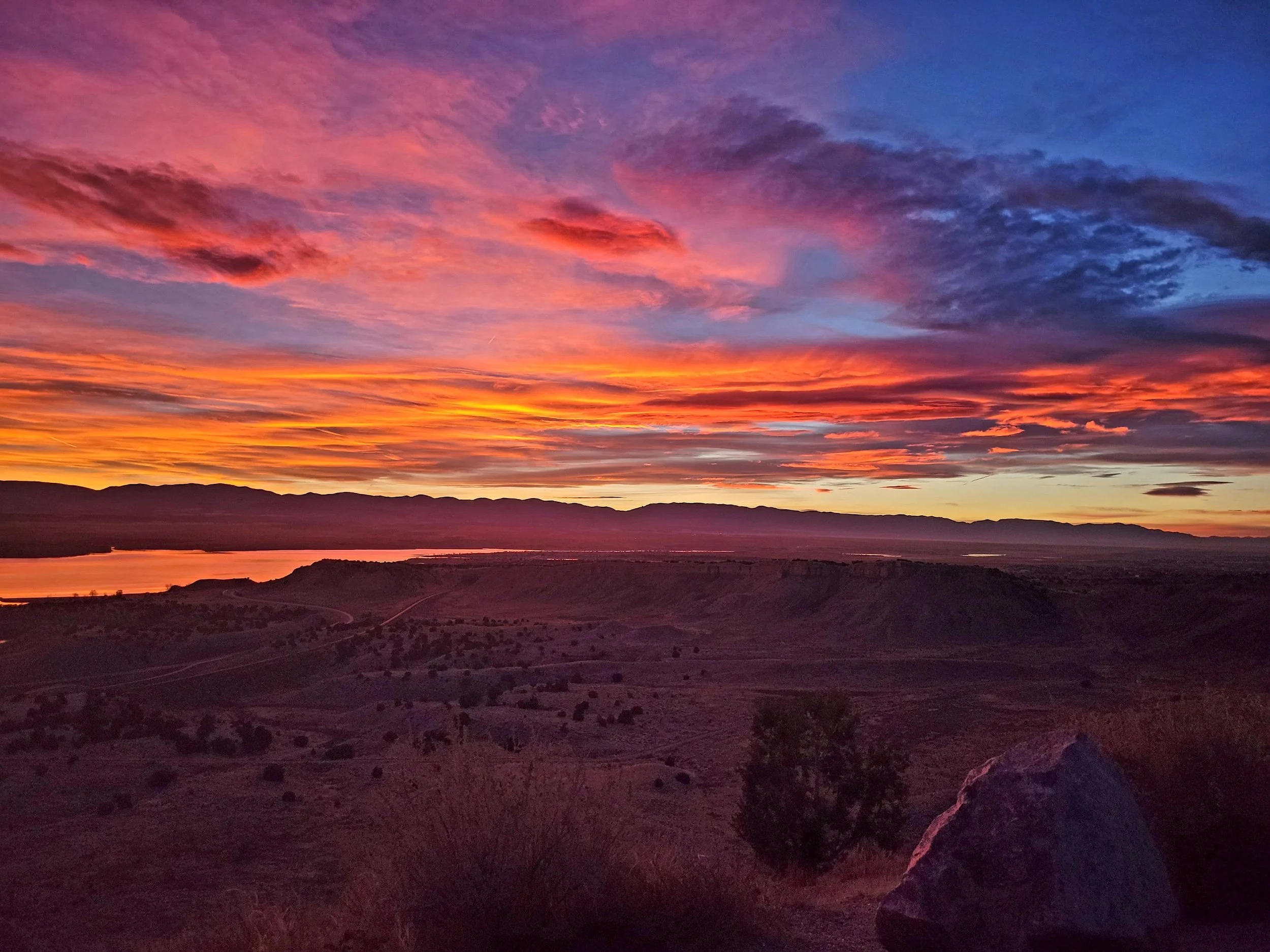 Colorful sunset over a landscape with hills and a body of water, with vibrant pink, orange, purple, and blue clouds in the sky in Pueblo West, Colorado.