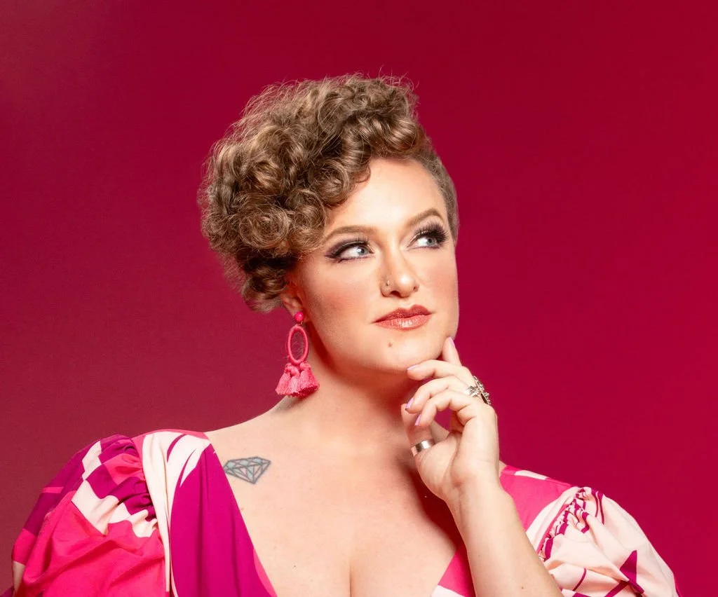 Freya West, a woman with brown, curly hair and makeup, wearing pink earrings and a pink, patterned dress, touching her chin with her hand against a pink background.