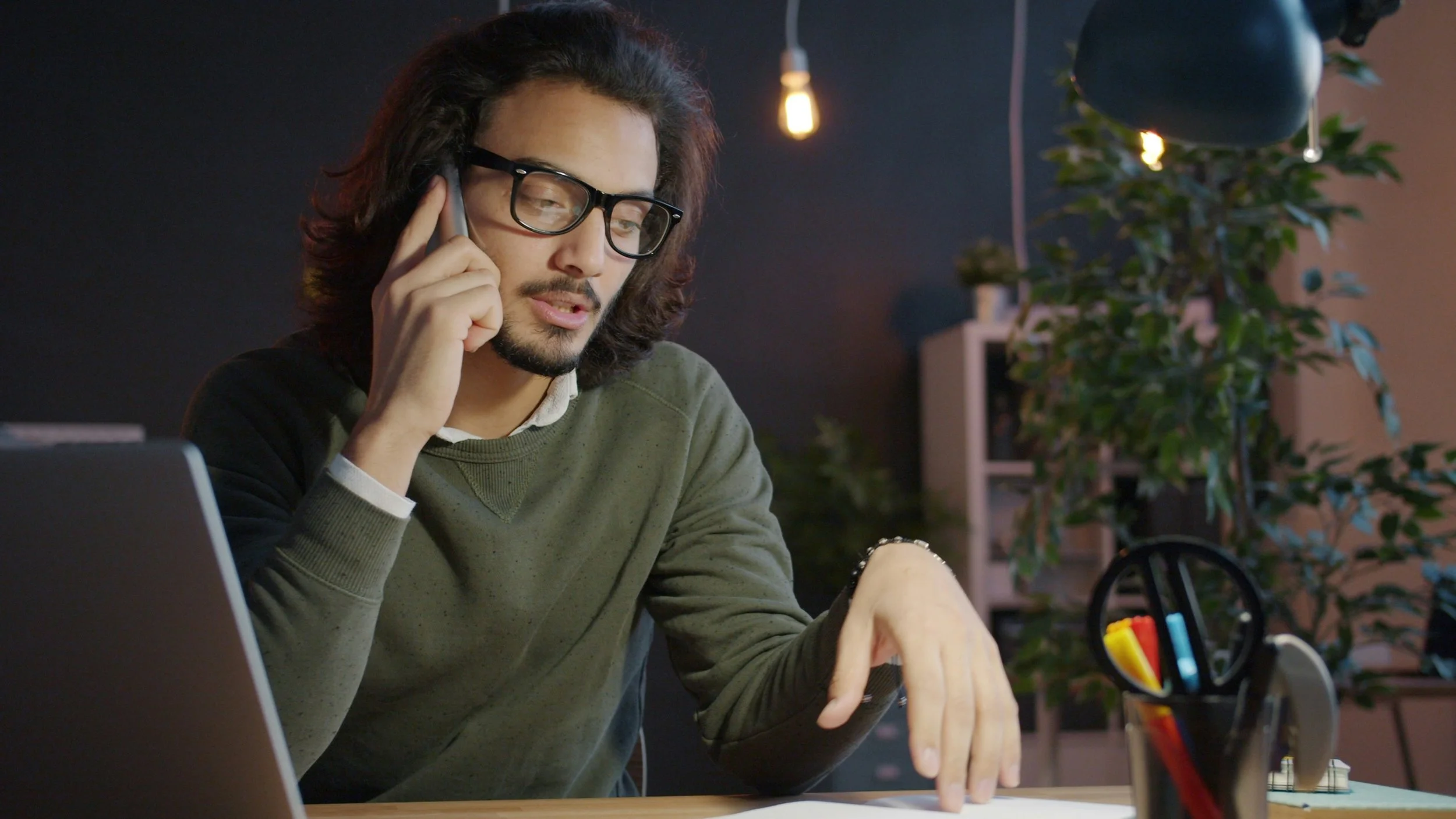 Man with long hair and glasses talking on a phone while working at a desk with office supplies, inside an office with plants and warm lighting.