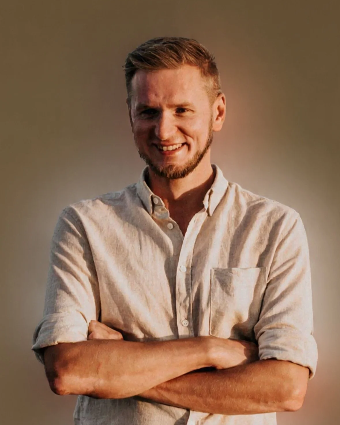 A young man with short, light-colored hair, a trimmed beard, and a smile, standing with arms crossed, wearing a white button-up shirt against a plain background.