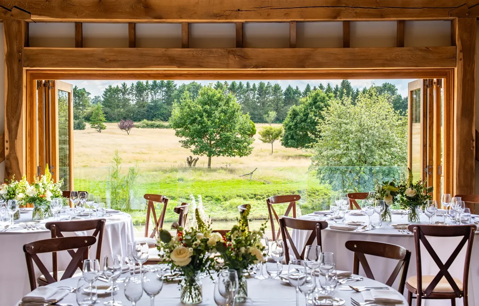 A rustic dining room with large open windows showcasing a scenic outdoor view of trees and grassy fields, decorated with round tables set with white tablecloths, glassware, and floral centerpieces.