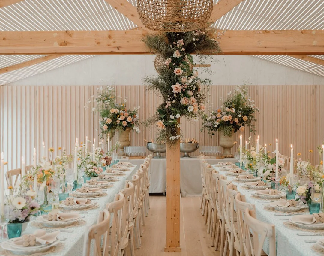Long banquet table decorated with floral arrangements, candles, and tableware in a rustic wooden pavilion.