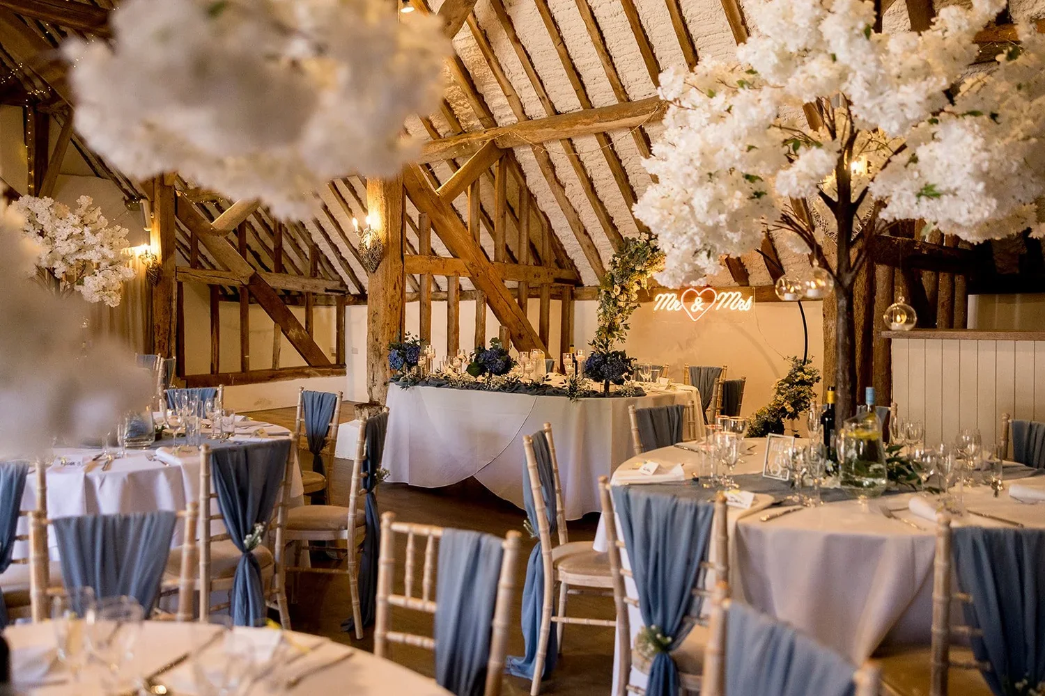 Wedding reception with decorated tables, floral arrangements, and a heart neon sign that reads "Mr & Mrs" on a rustic wooden ceiling.
