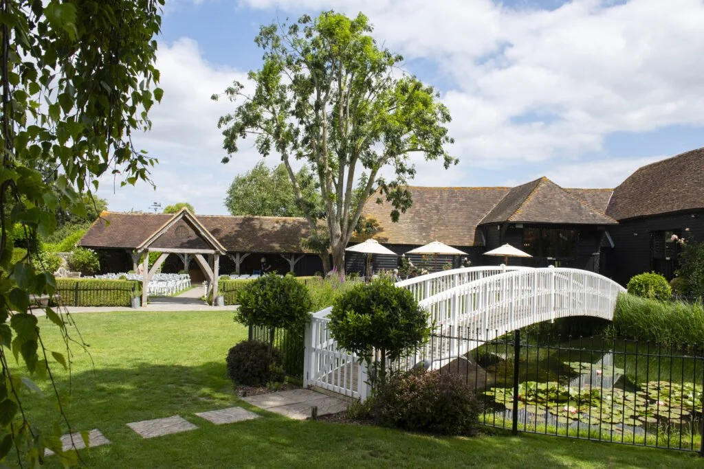 A dog-friendly wedding venue showing a garden with a pond, white bridge, wooden buildings, and a gazebo under a blue sky with clouds.