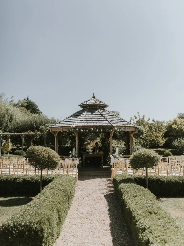 Outdoor dog-friendly wedding ceremony setup with a gazebo, rows of chairs, and neatly trimmed hedges and trees under a clear sky.