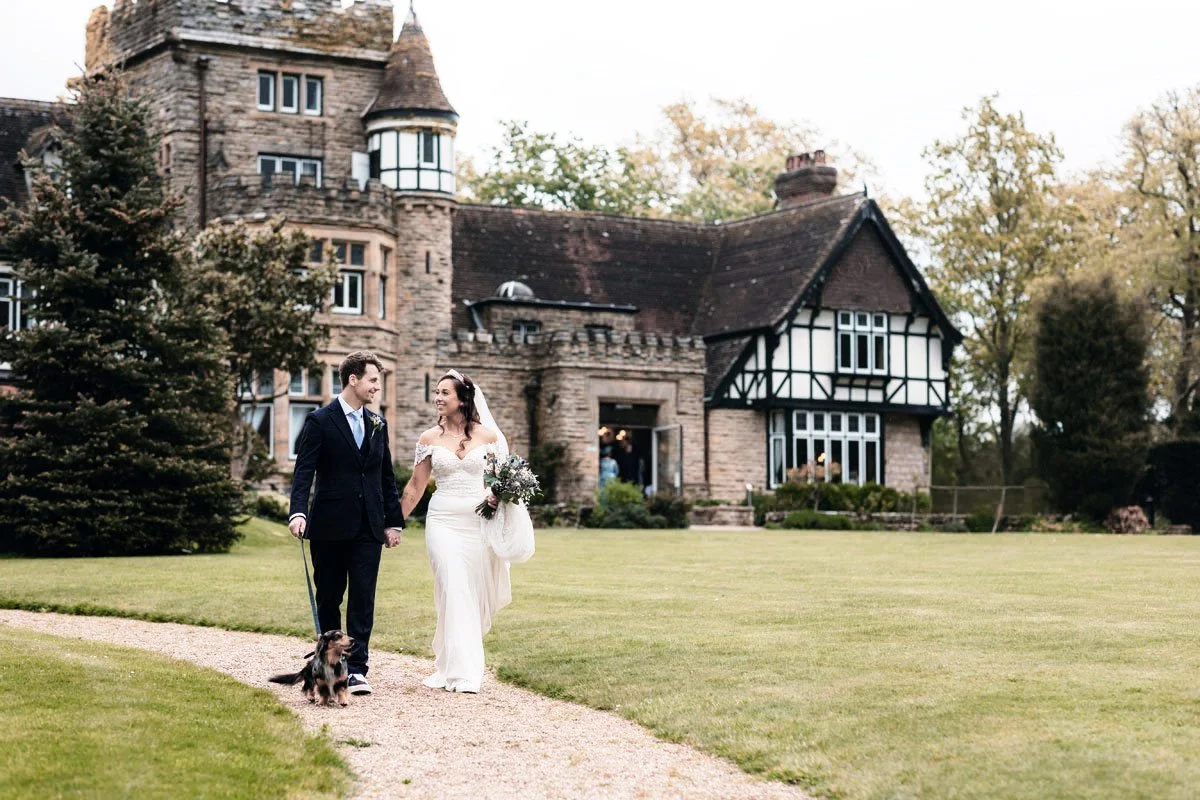 Bride and groom walking a dog on a gravel path in front of a large historic house, wedding day, casual and happy moment.