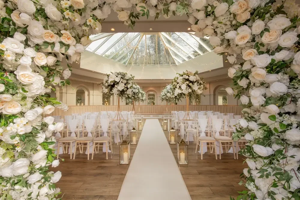 Elegant wedding ceremony setup with white floral arch and aisle, floral arrangements on tall stands, and white chairs inside a glass-roofed venue.
