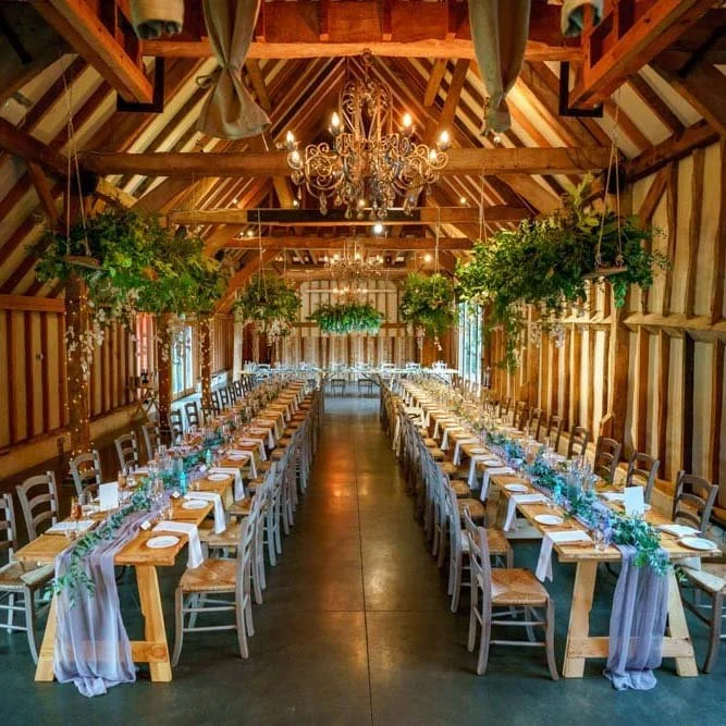 Long rectangular wooden tables set for a wedding reception inside a rustic barn, decorated with greenery and fairy lights, with chandeliers hanging from the ceiling.