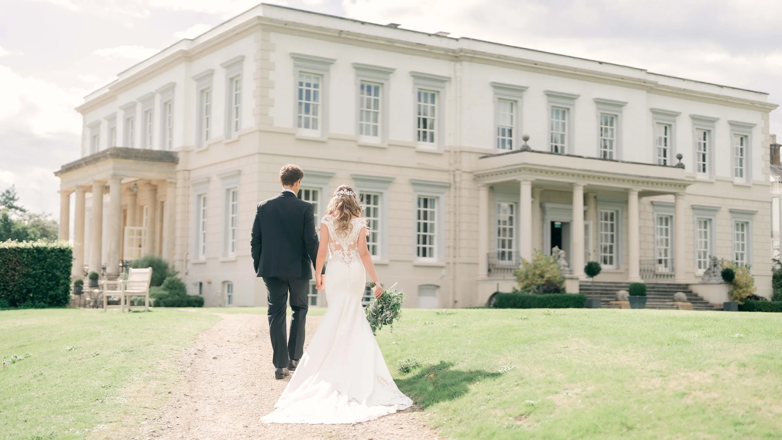 A bride and groom walking hand in hand towards a large white mansion on a sunny day.