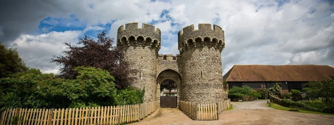 A medieval stone castle with two round towers and a gate, surrounded by a wooden fence and greenery under a cloudy sky.