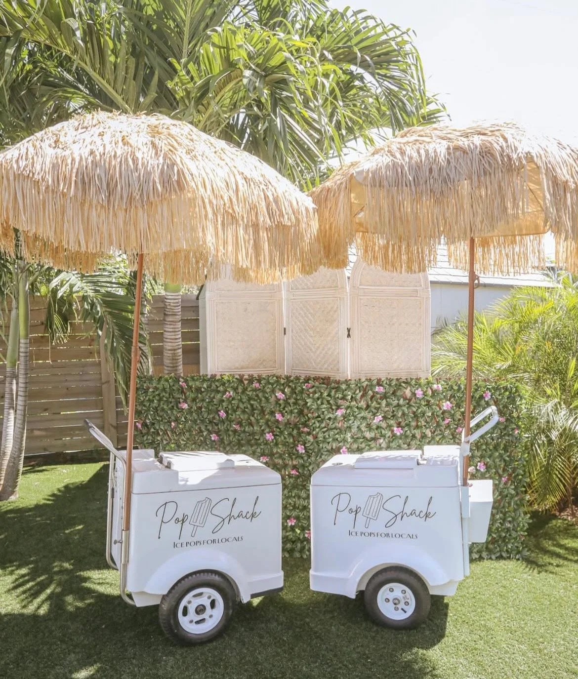 Two white ice cream carts labeled "Pop Shack" with a graphics of ice cream bars, set under two thatched umbrellas in a garden with tropical plants and pink flowers.