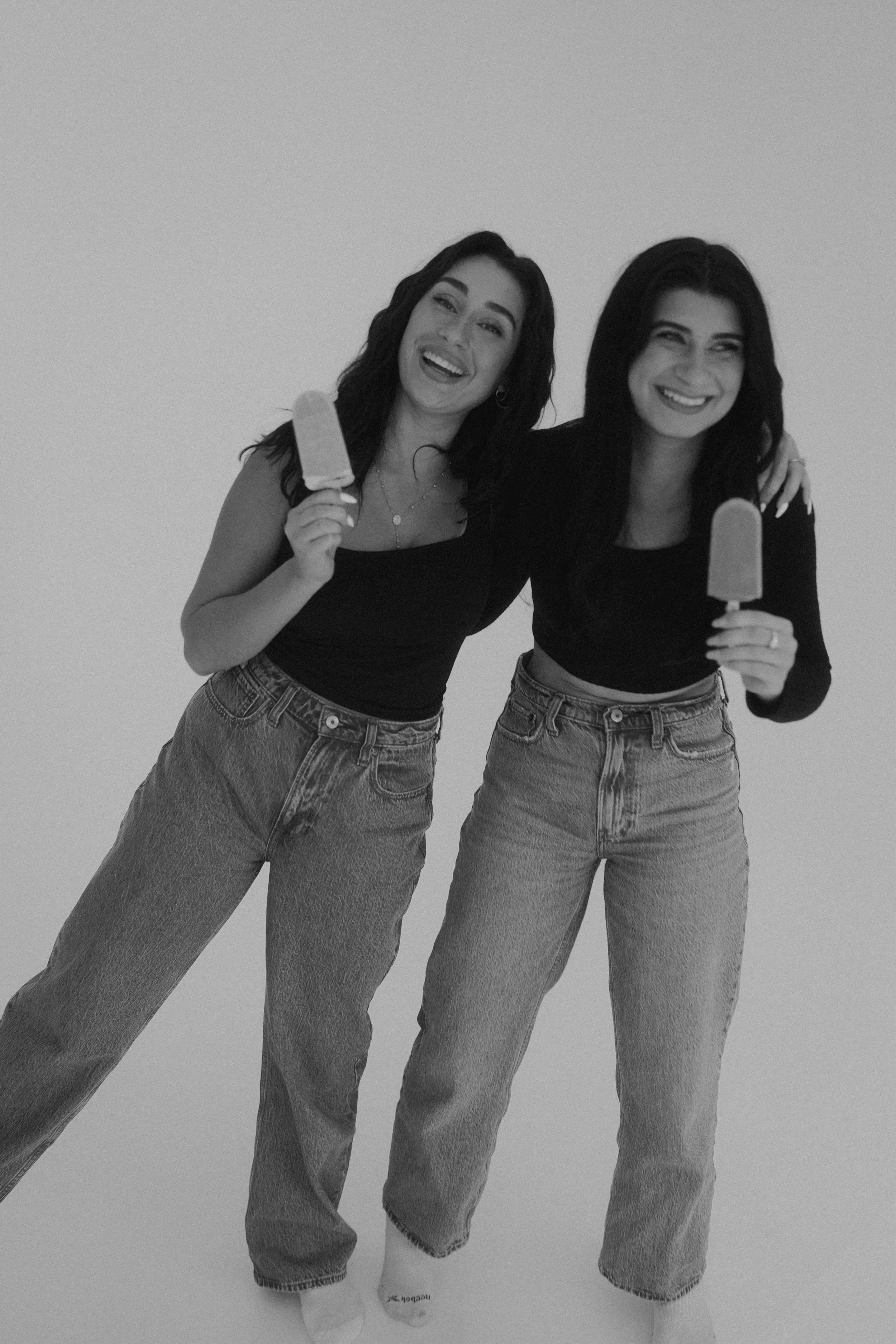 Two women standing side by side, smiling, each holding a popsicle, wearing black tops and jeans.