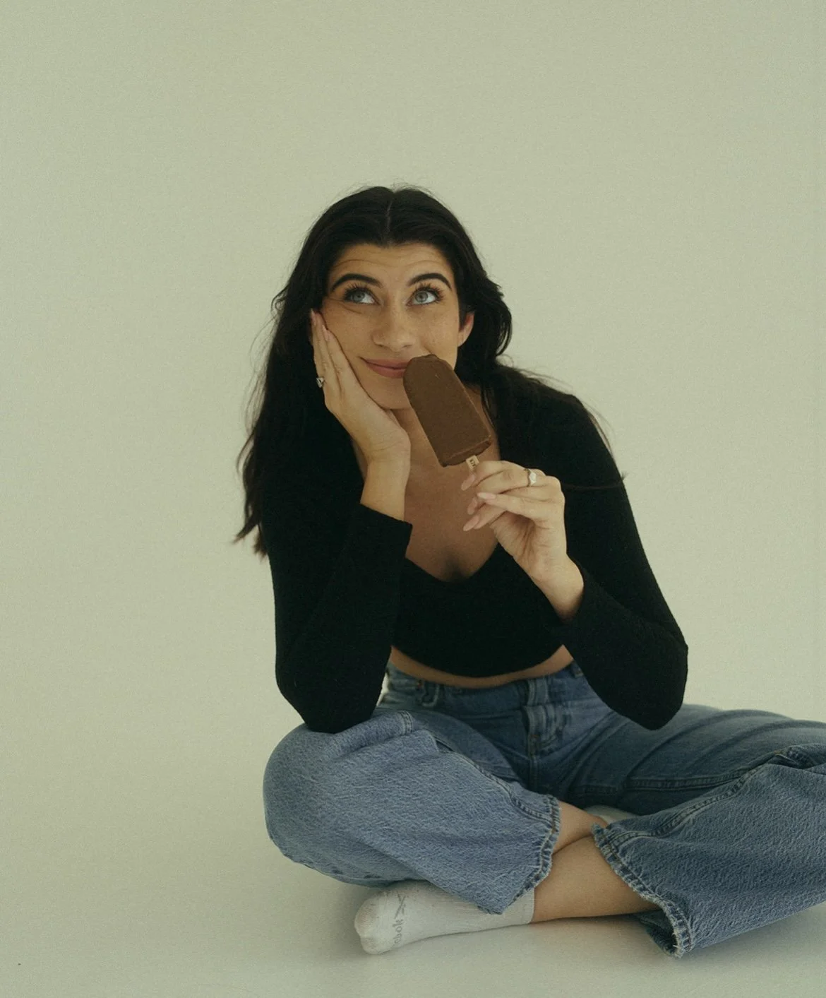 A woman sitting cross-legged on the floor, holding a chocolate popsicle, gazing upwards with a dreamy expression.