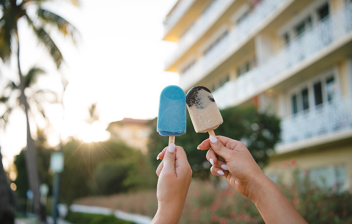 Two hands holding ice pops outdoors with a building and trees in the background, sunlight shining.