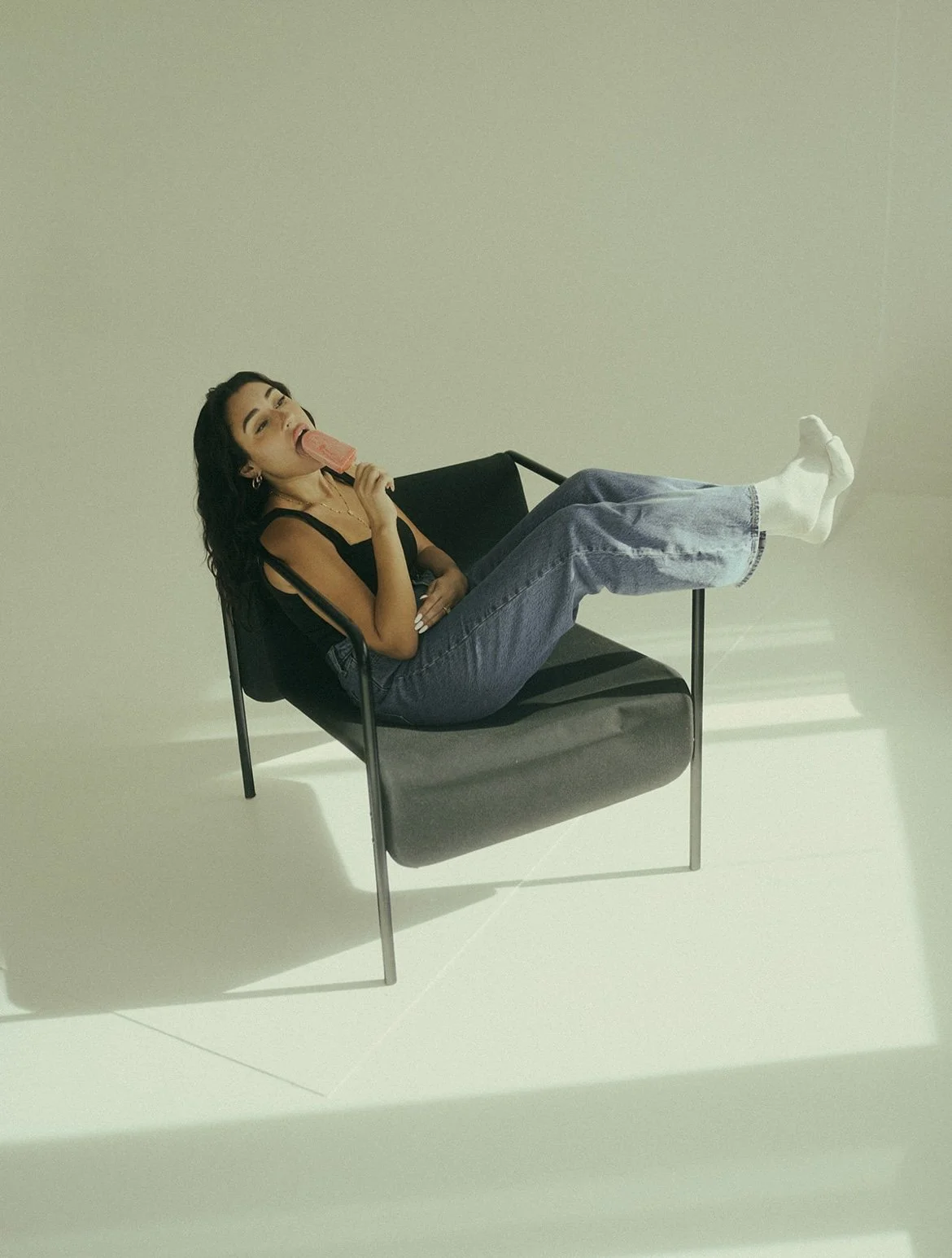 Young woman with dark hair, wearing a black top and jeans, sitting on a modern black chair, and licking a pink popsicle.