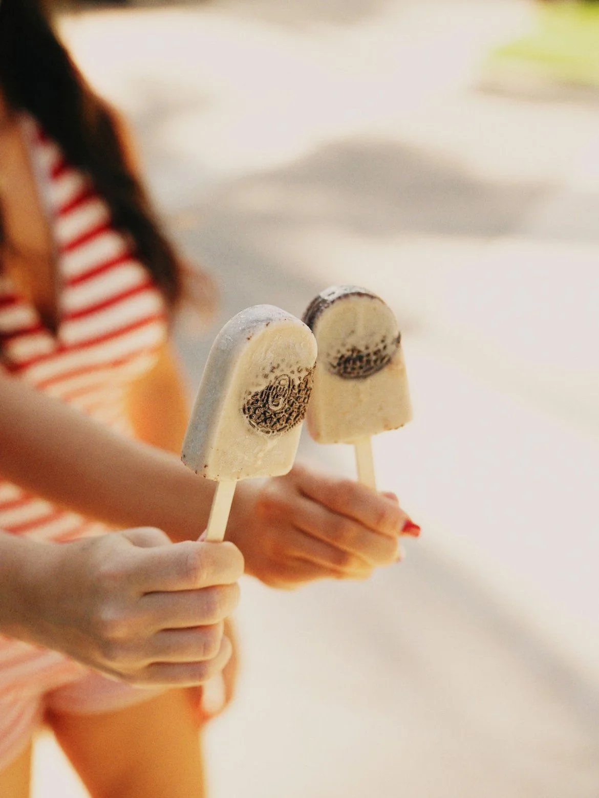 Close-up of a person holding two vanilla ice cream popsicles with Oreo cookies in a sunny outdoor setting.