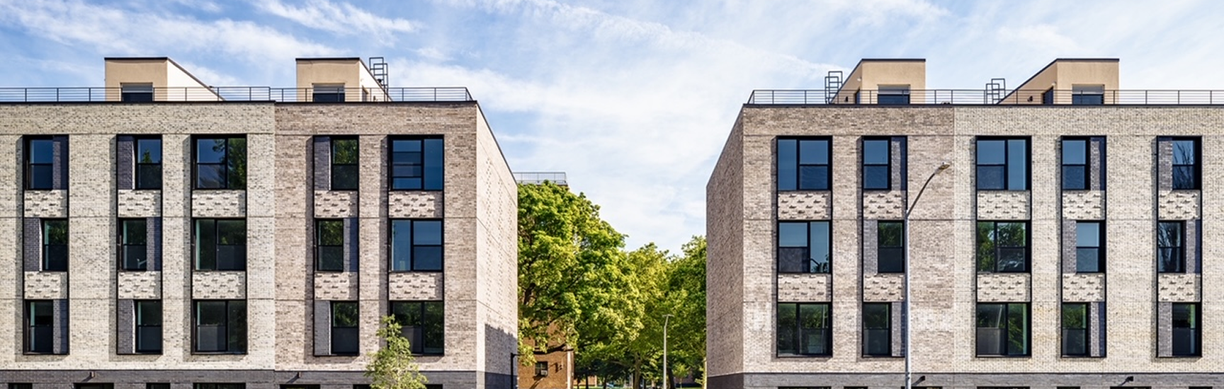 Modern residential apartment buildings with brick facades, multiple windows, and rooftop terraces, flanked by trees under a partly cloudy sky.