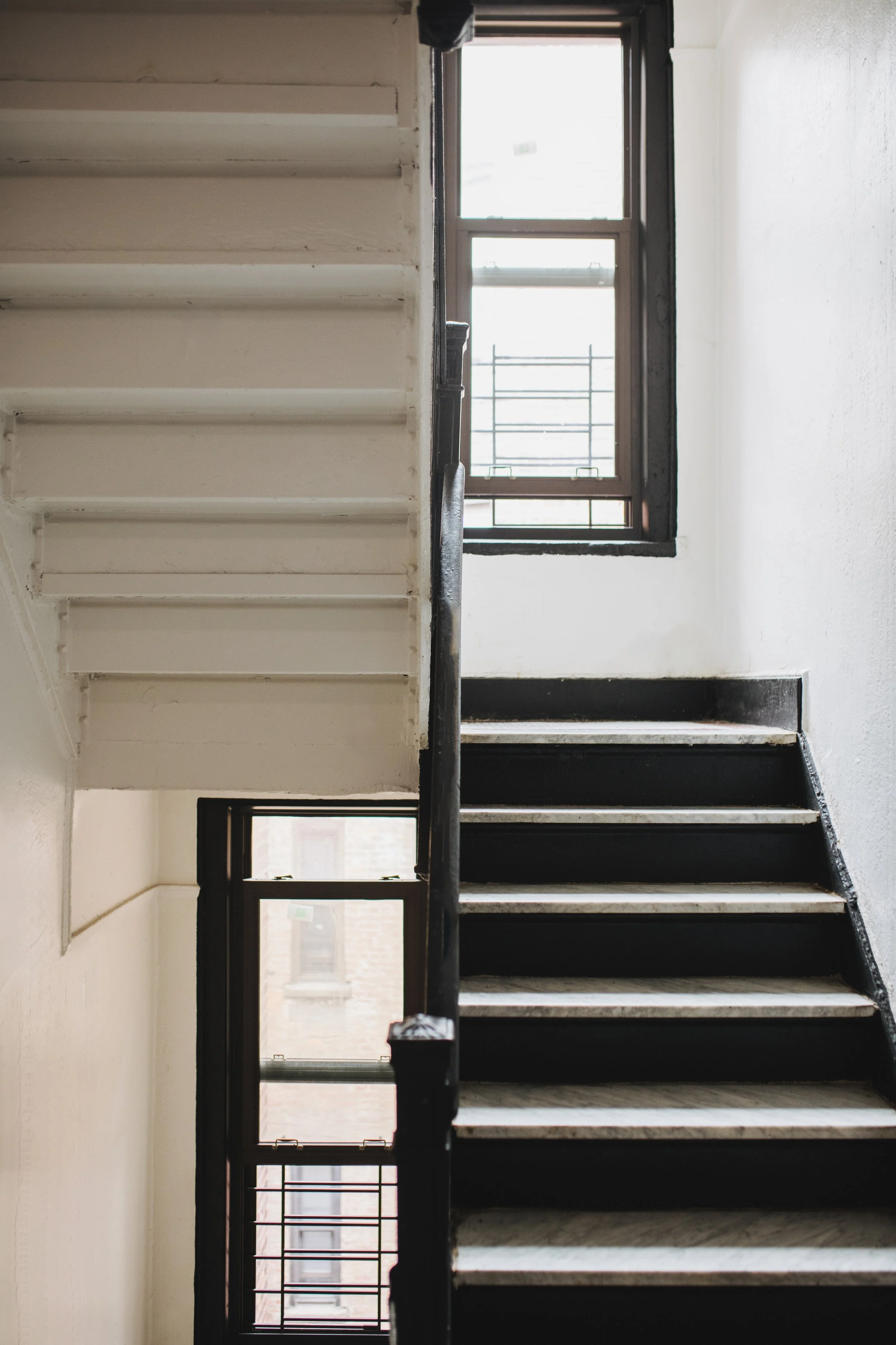Interior view of a staircase with black steps and a black handrail, next to a wall with large windows letting in natural light.