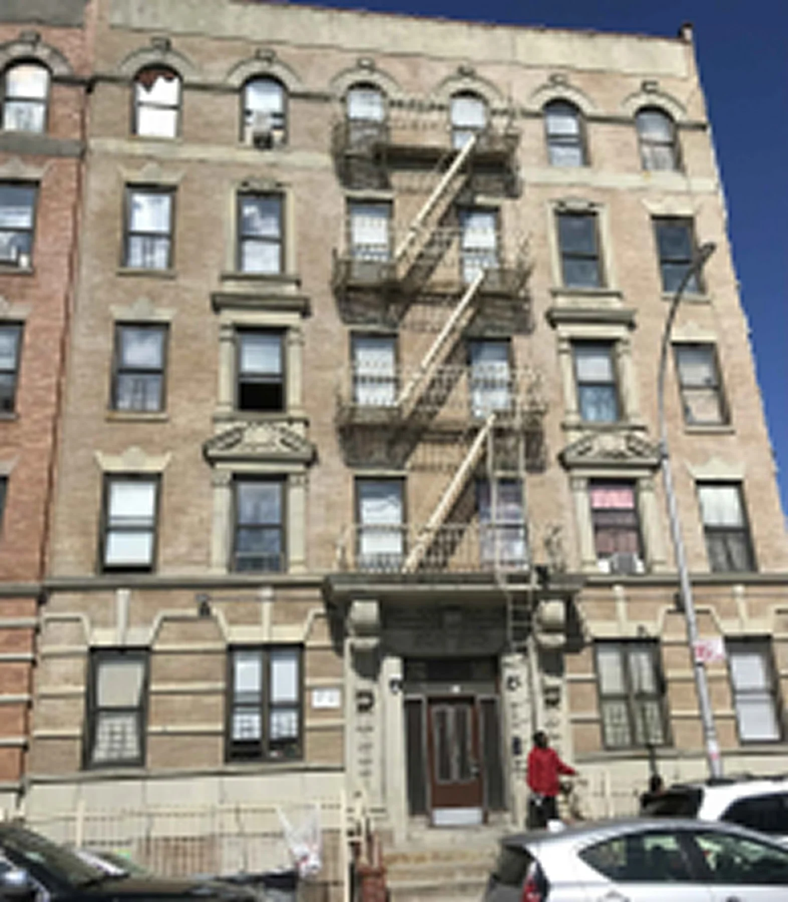 A multi-story brick apartment building with fire escapes on the front and a person in a red jacket walking by, parked cars, and a streetlamp in the foreground.