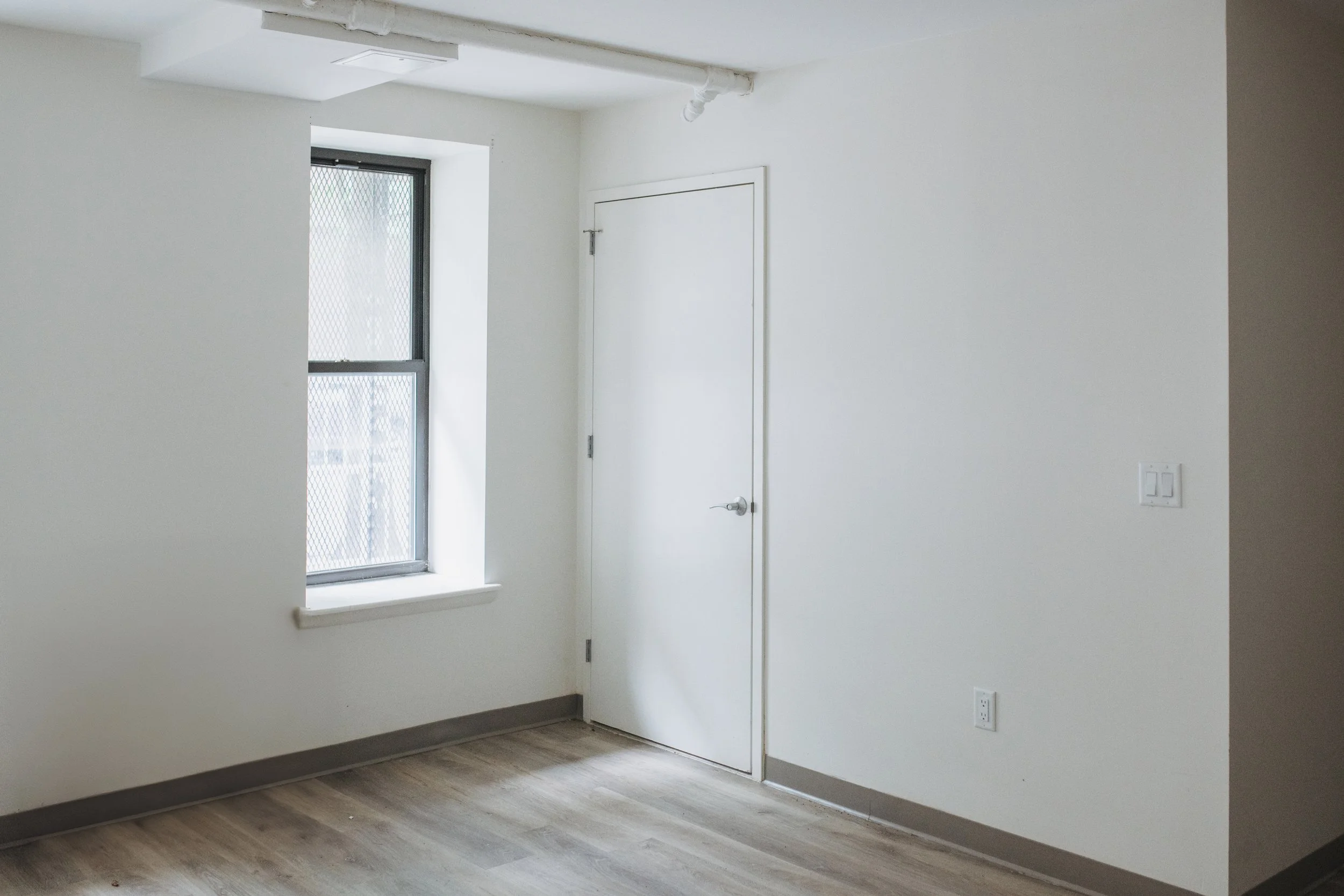 Empty room with white walls, a window, and a closed door, with laminate flooring and electrical outlets.
