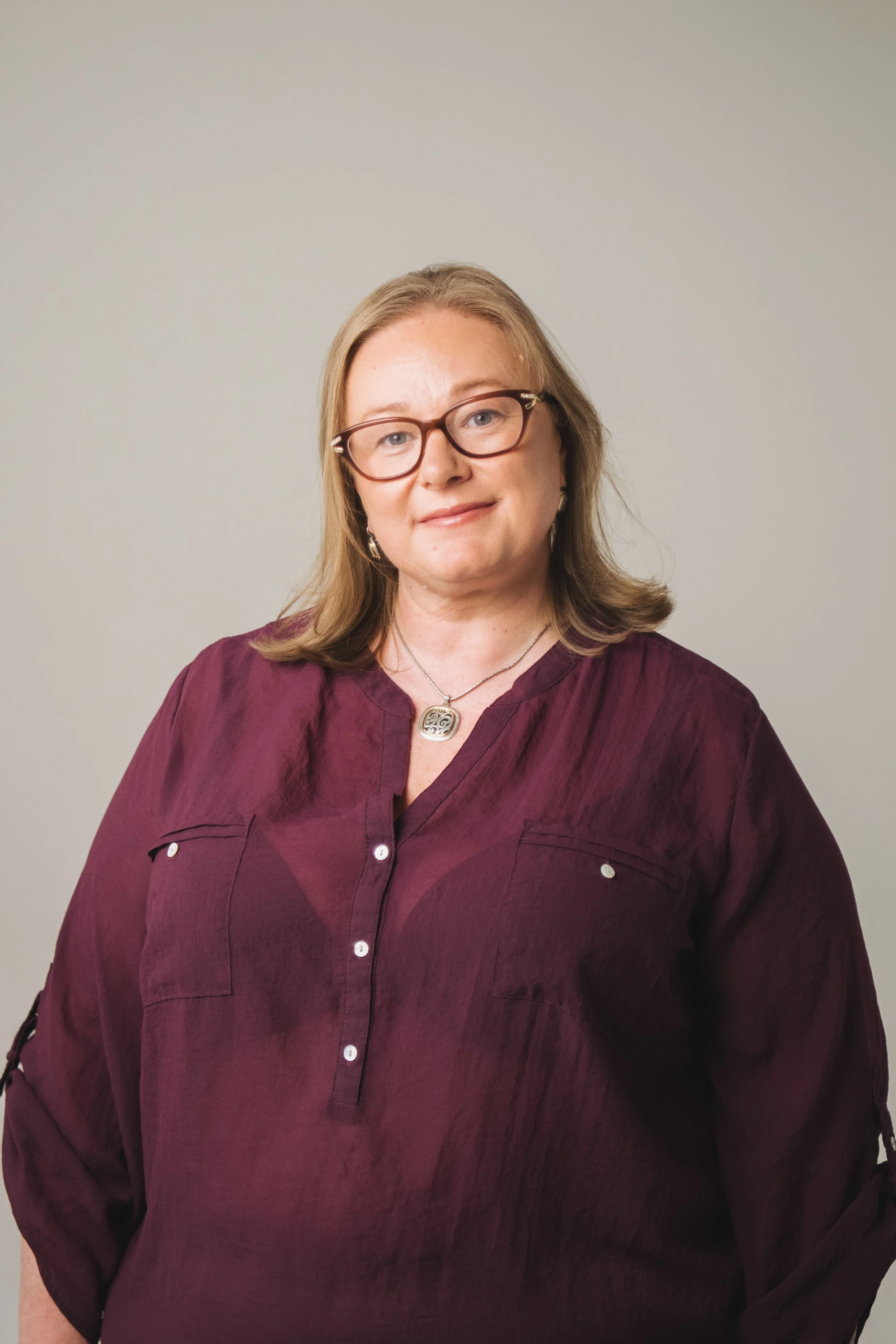 A woman with shoulder-length blond hair, glasses, and a necklace stands against a plain light-colored wall, wearing a dark red button-up blouse.