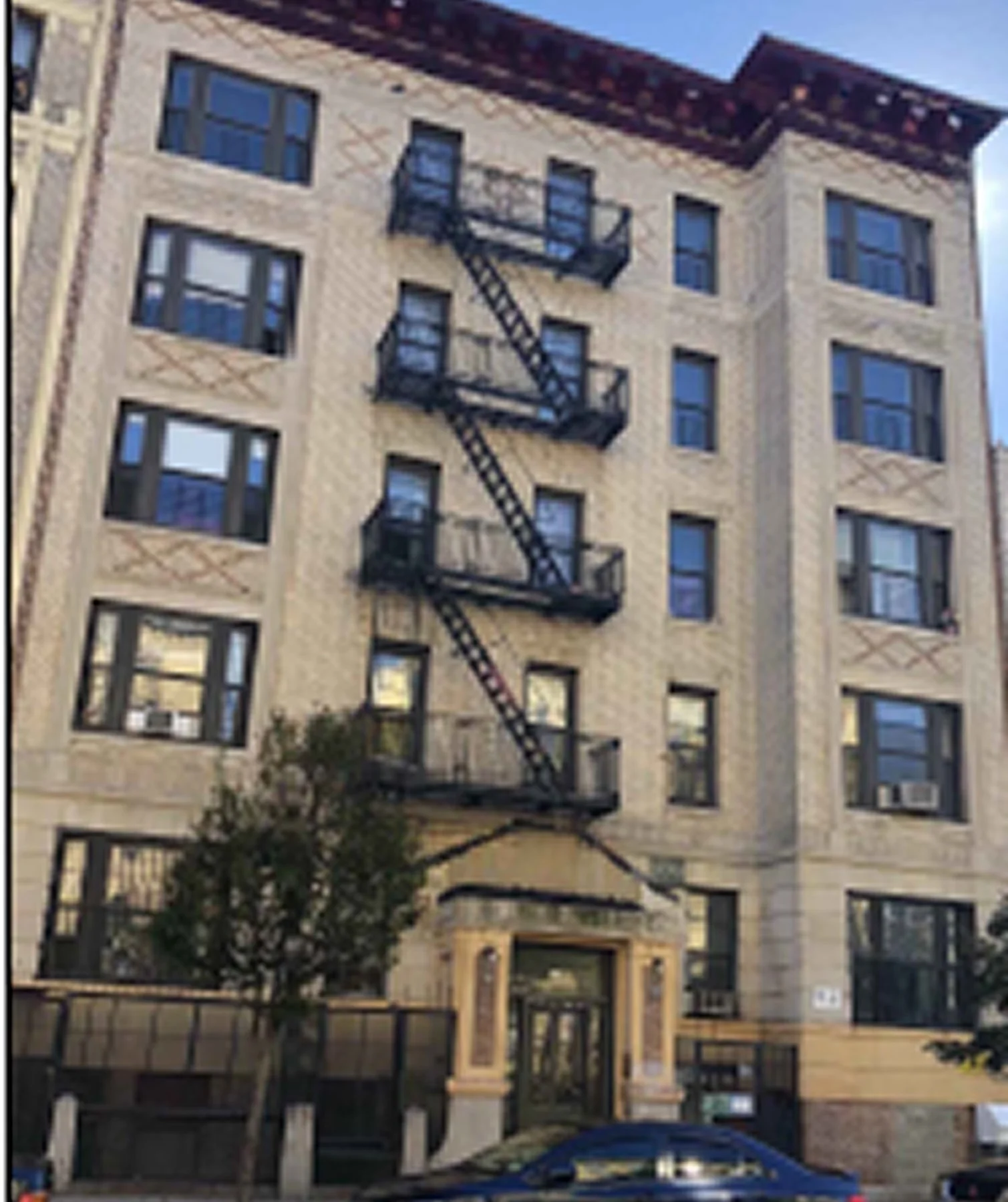 A multi-story residential building with beige brick exterior, multiple black fire escape balconies, several windows, and a small tree in front. The building appears to be under construction or renovation, with an unfinished wooden frame near the entr