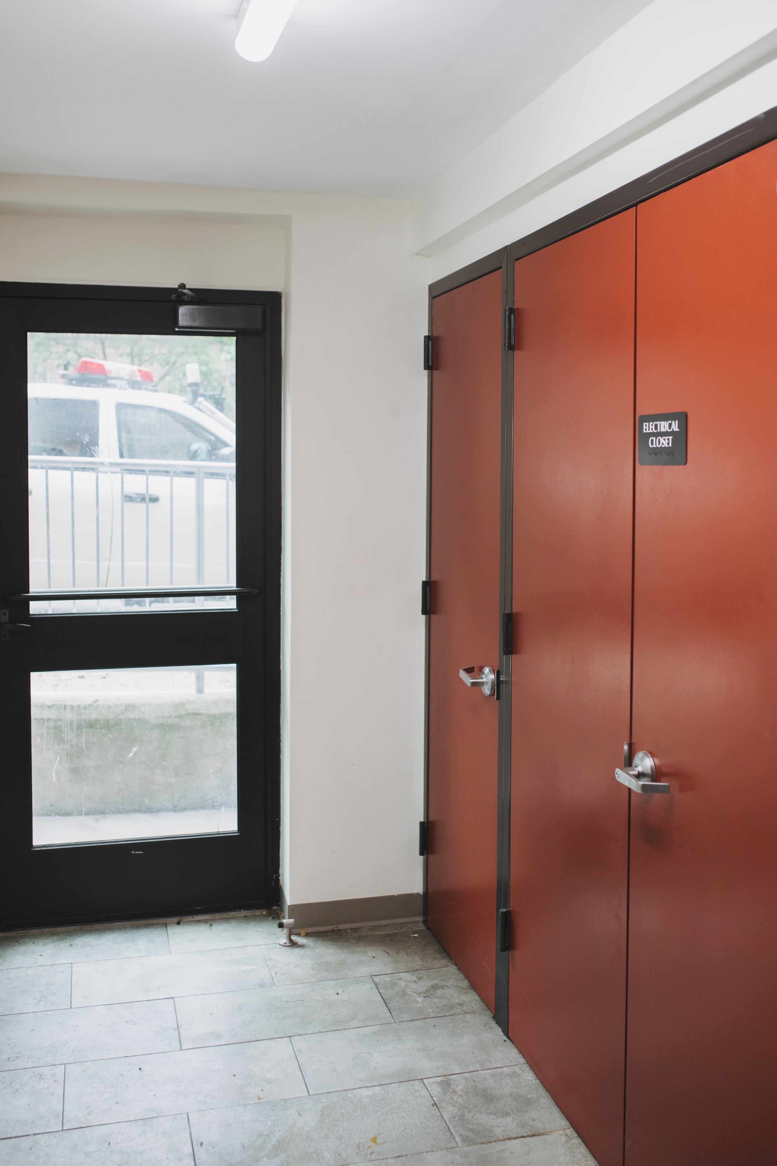 Inside view of a red electrical closet door with a silver handle and a black sign that reads 'Electrical Closet.' Next to it is a black exit door with a push bar and a window, through which a police vehicle is visible outside.