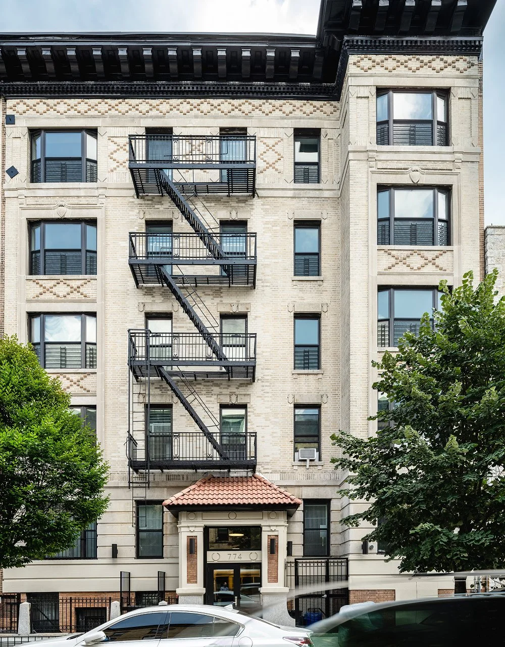 A multi-story residential building with a brick facade, multiple windows with black frames, and black fire escape stairs on the front. Green trees are in front of the building, and a white car is parked near the entrance.