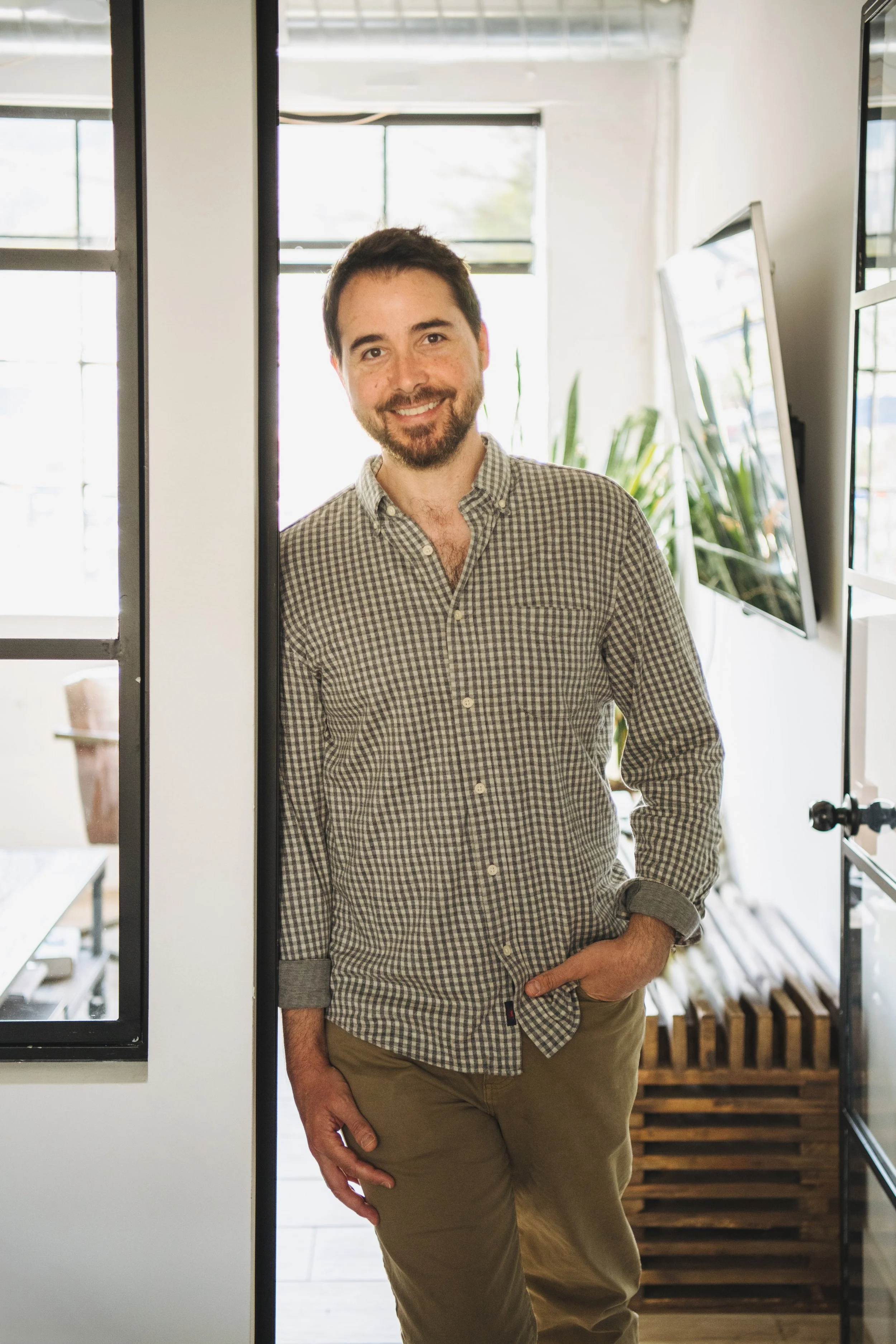 A man with dark hair and a beard, wearing a checkered shirt and beige pants, standing in a modern, bright interior space with large windows and plants, smiling at the camera.