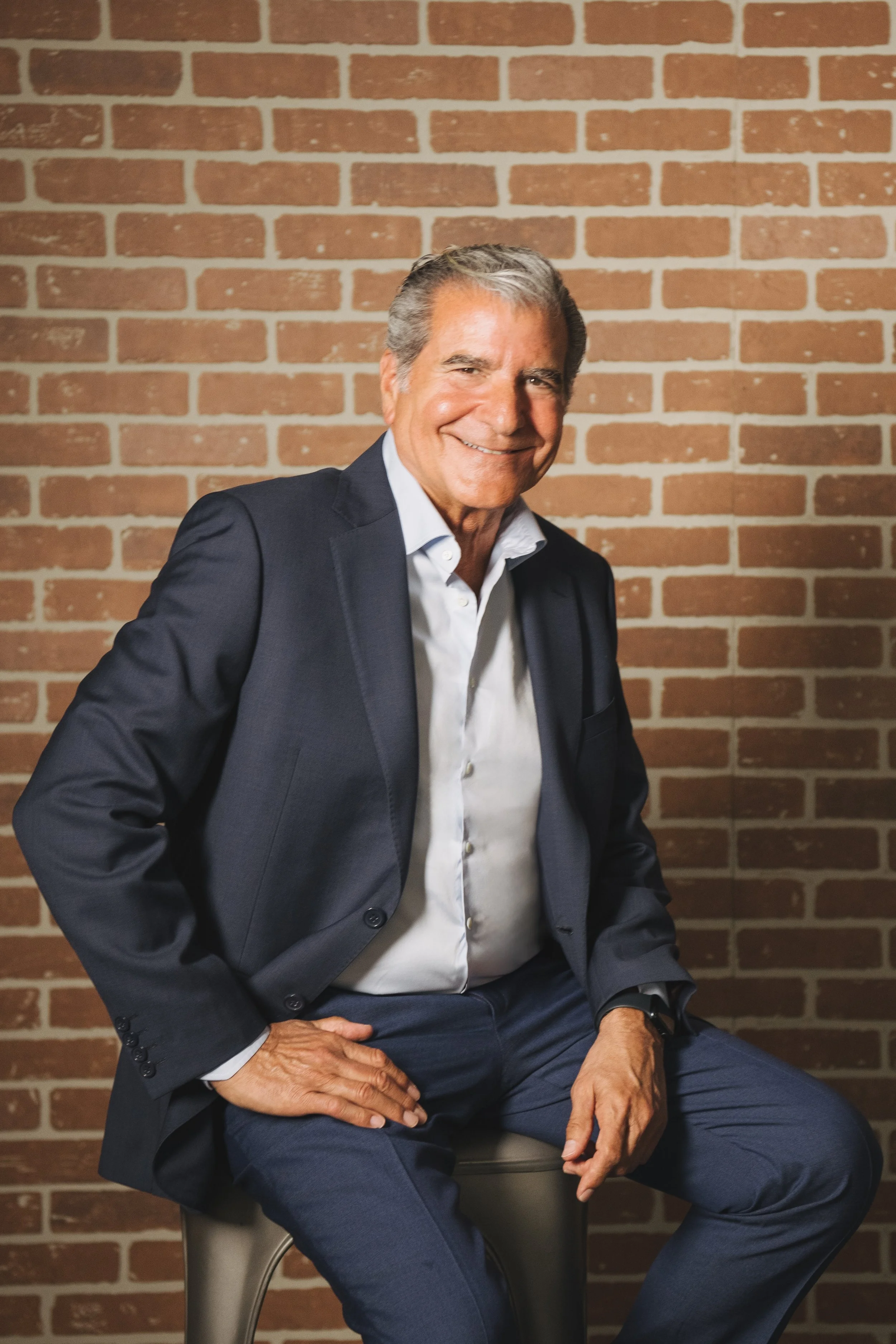 A smiling older man in a business suit sitting on a chair against a brick wall, with one hand on his lap and the other on his knee.
