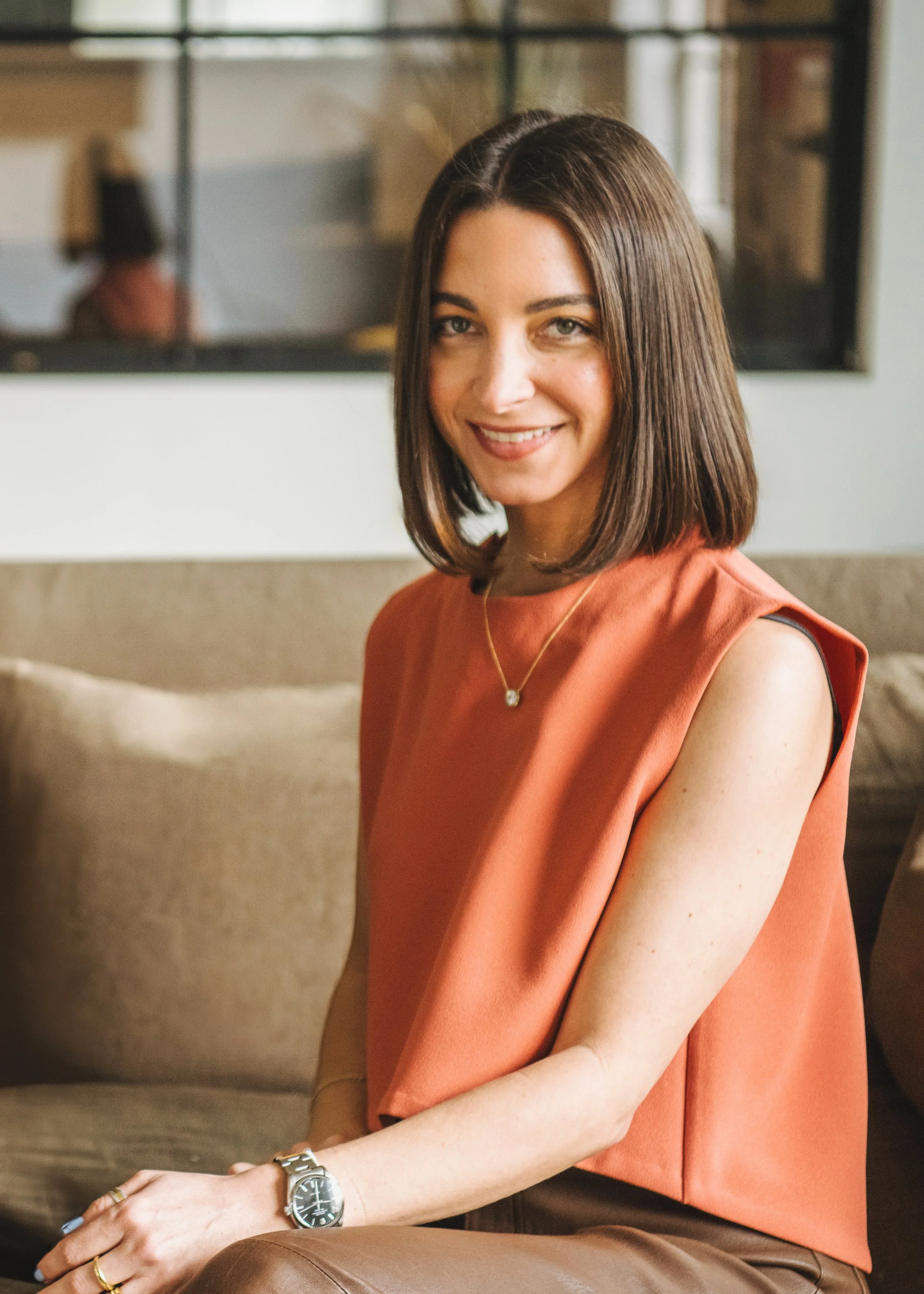 A woman with shoulder-length brown hair, wearing a sleeveless orange top, sitting on a wooden surface with a black metal frame, smiling at the camera, with her hands resting on her lap, wearing a wristwatch and rings.