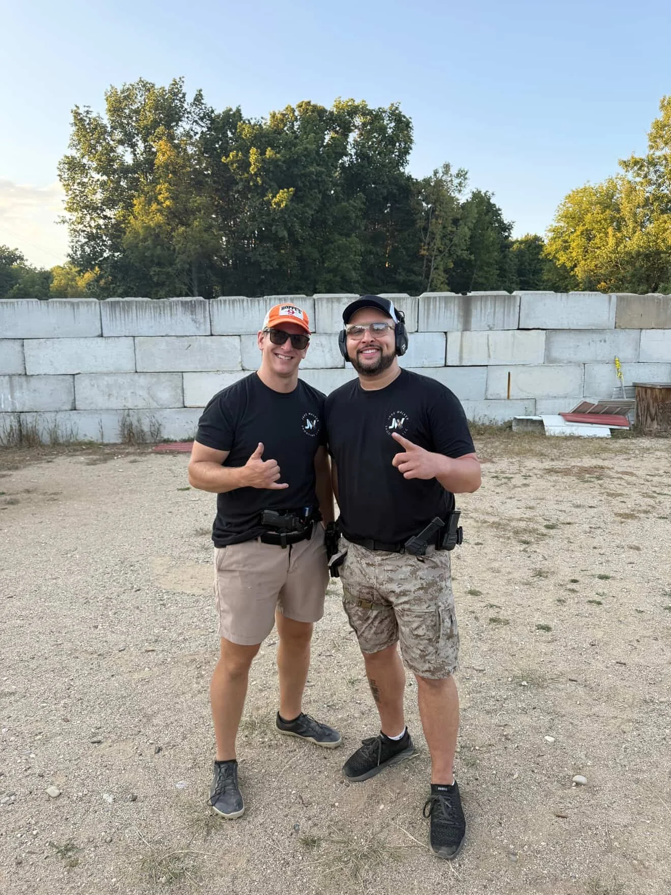 Two men at an outdoor shooting range, posing and smiling at the camera. One gives a thumbs-up and the other points with his finger. They are wearing safety glasses and casual clothing, with the man on the right also wearing hearing protection.