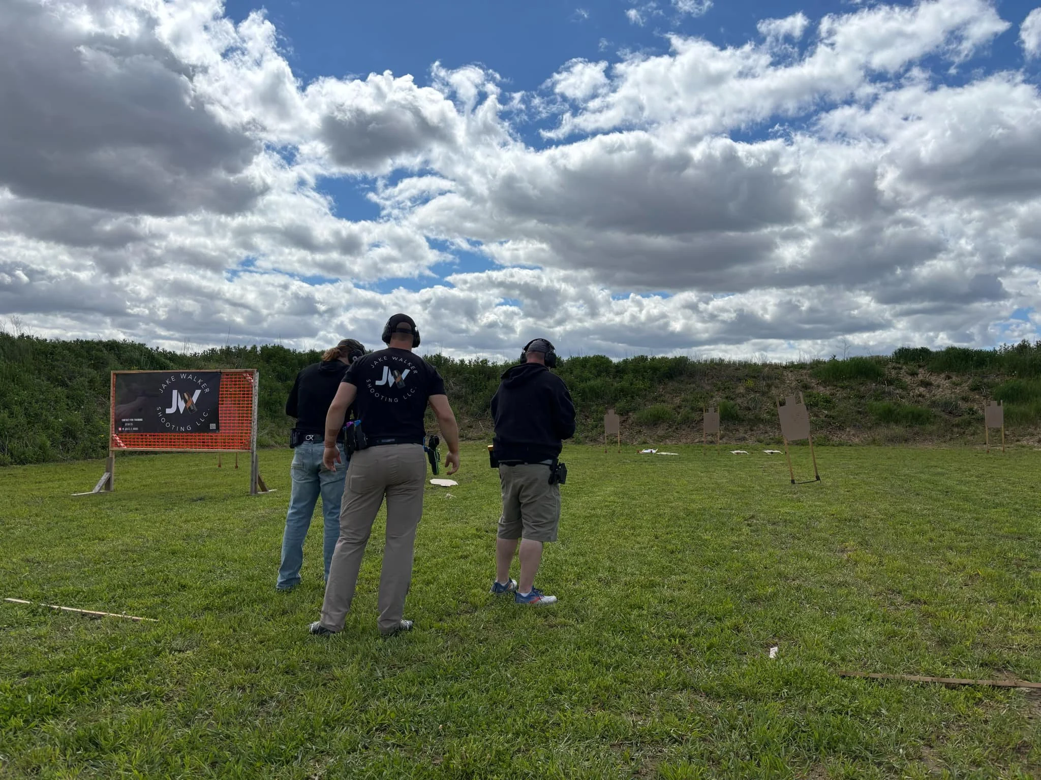 Three people standing on a grassy field at a shooting range, with targets set up in the distance and a cloudy sky overhead.