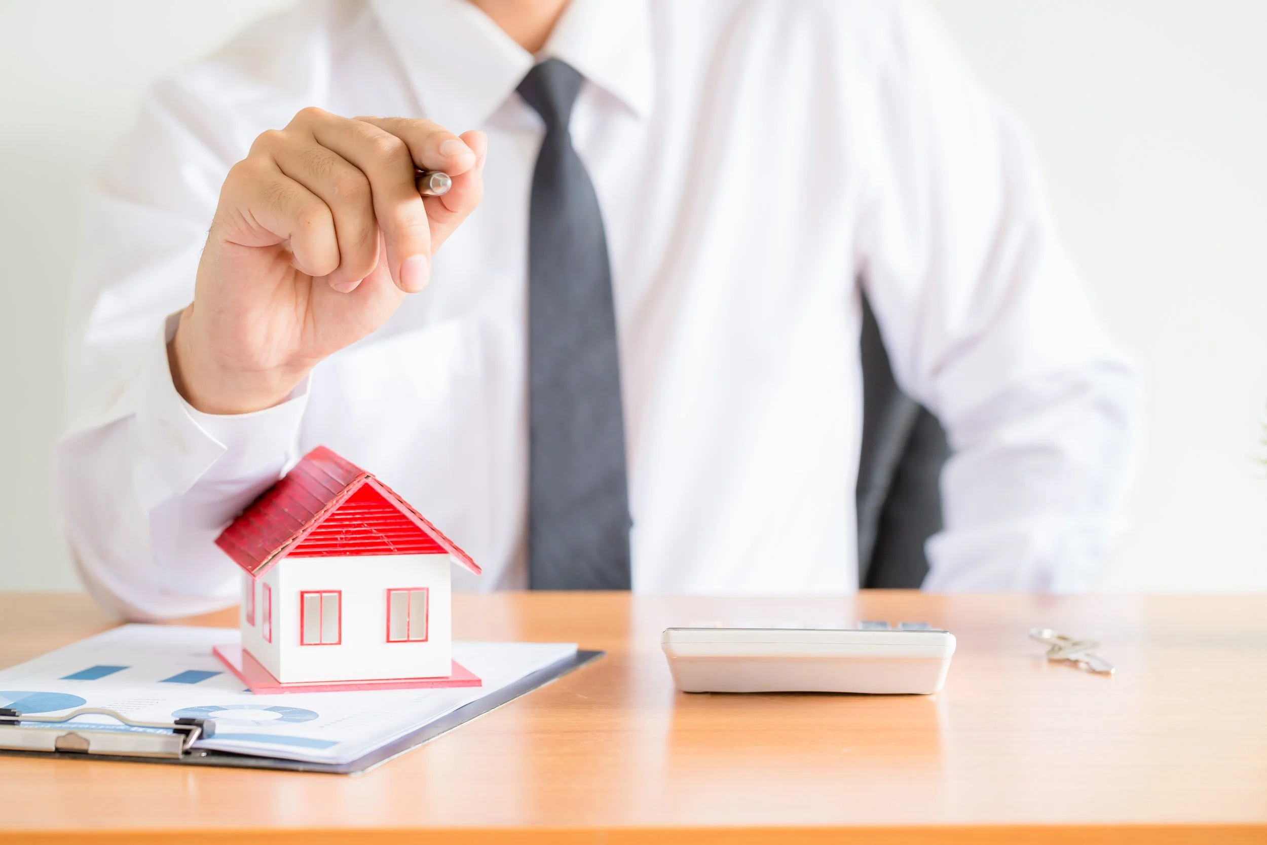 A person in a white shirt and black tie sitting at a desk, holding a pen above a small model house with a red roof, along with keys, a clipboard, and a calculator on the desk.