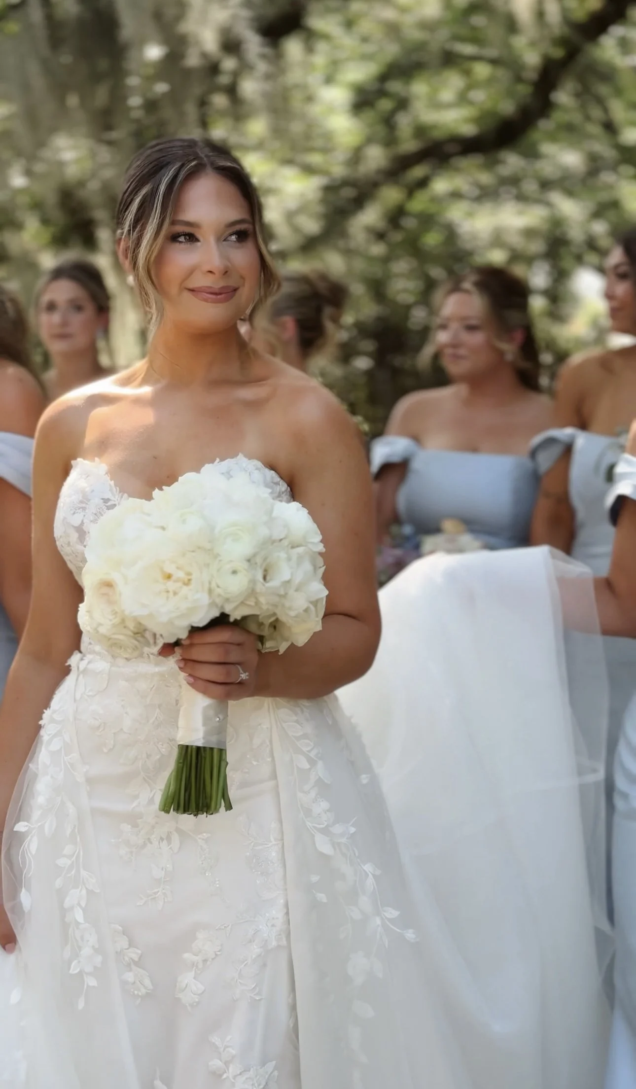 A bride in a white wedding dress holding a bouquet of white flowers, standing outdoors with bridesmaids in light blue dresses in the background. Wedding Content Creator, Louisiana Wedding Content Creator, New Orleans Wedding Content Creator,