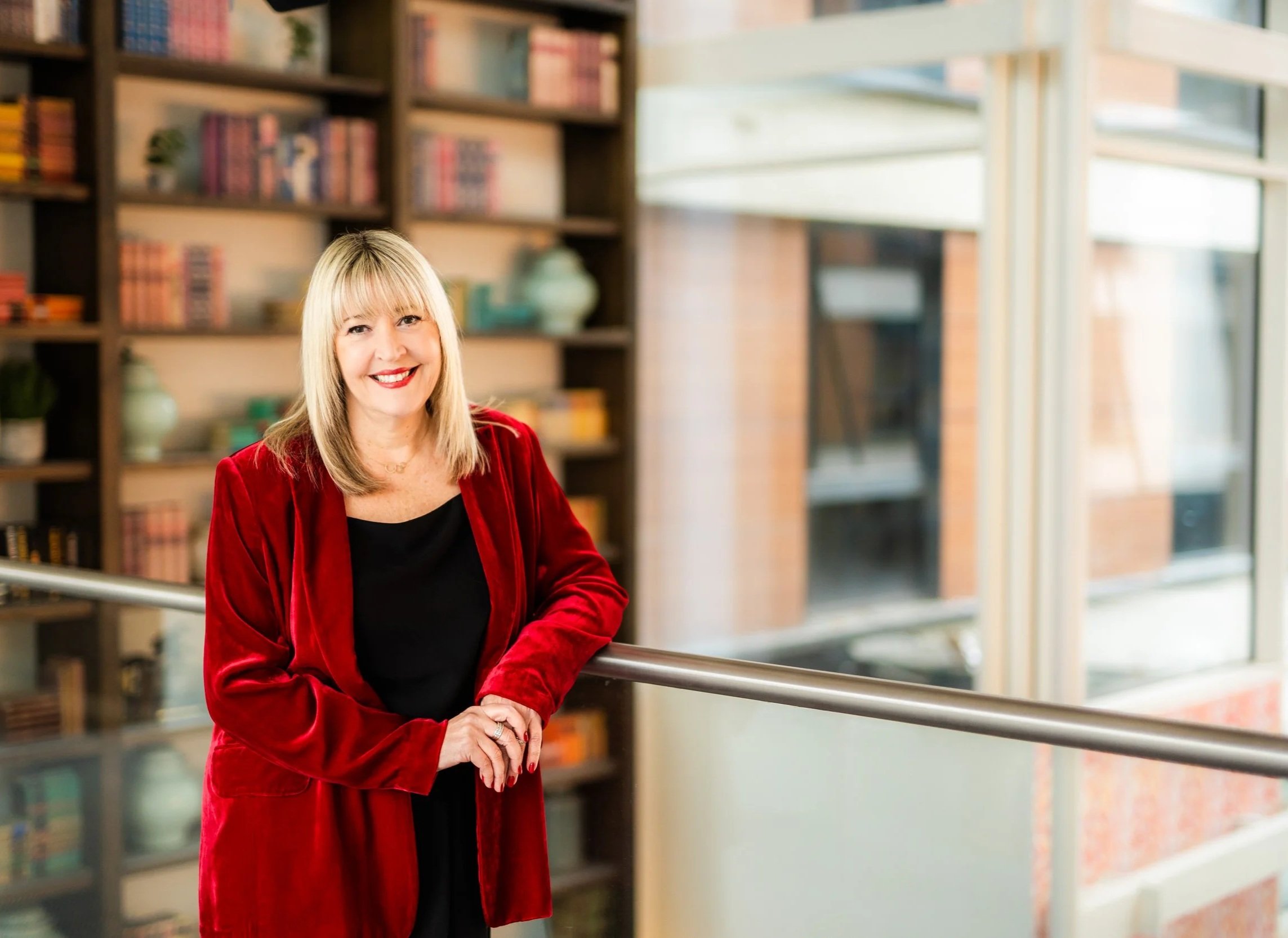 A woman with blonde hair, wearing a red velvet blazer and black top, smiling and leaning on a metal railing inside a building with a wood bookshelf filled with books and decorative items in the background, and large windows letting in natural light.