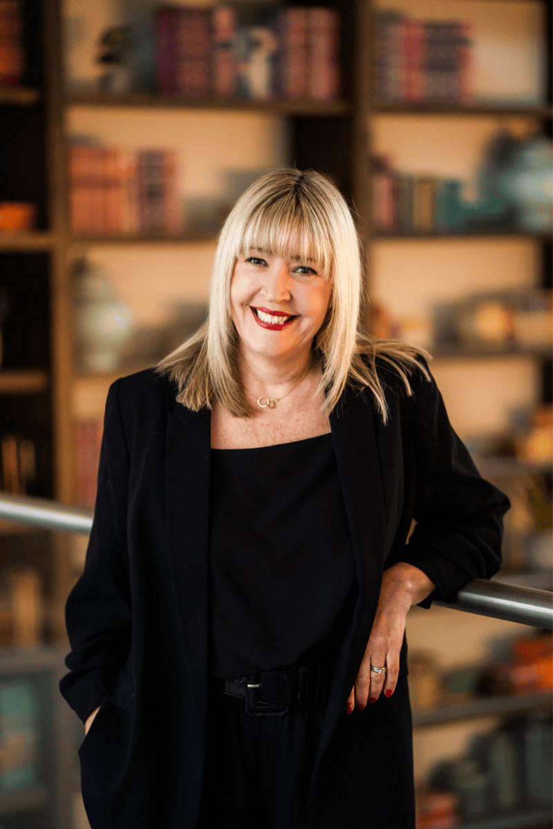 Bestselling author Lisa Timoney, with blonde hair and red lipstick wearing a black blazer and top, standing indoors in front of bookshelves filled with books.