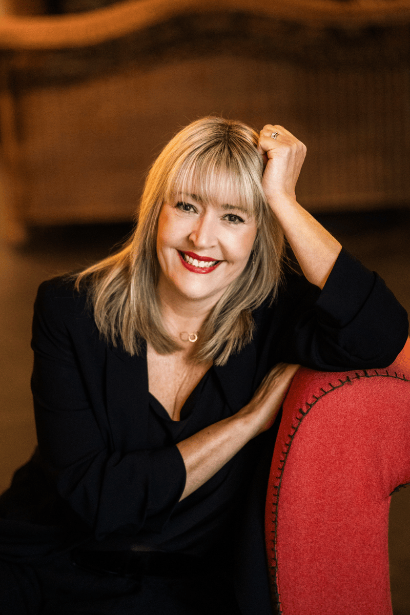 Bestselling author Lisa Timoney,  with blonde hair smiling, resting her head on a red chair, wearing a black top, in an indoor setting.
