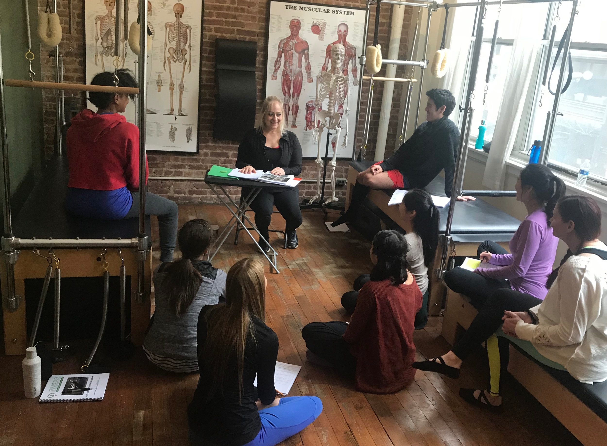A group of women sitting and listening to a woman teaching in a Pilates studio with anatomical posters and Pilates equipment.