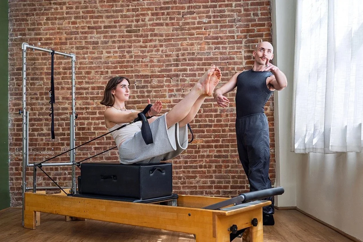 A woman is practicing Pilates on a reformer machine with the guidance of an instructor in a studio with exposed brick walls and anatomical posters.