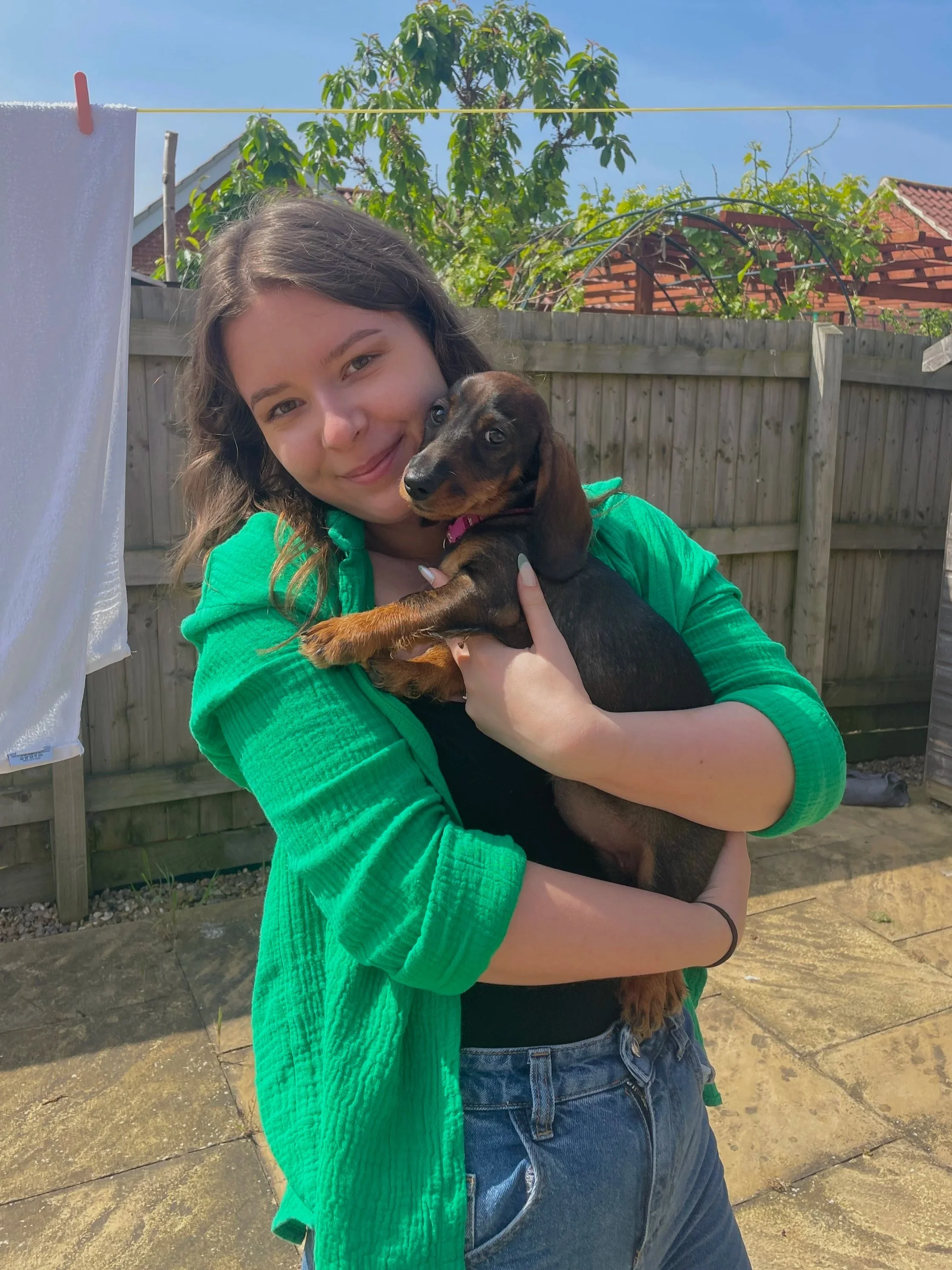 A young woman with long, wavy brown hair holding a small brown and black puppy outdoors on a sunny day in a backyard, with a wooden fence, green trees, and a clothesline in the background.