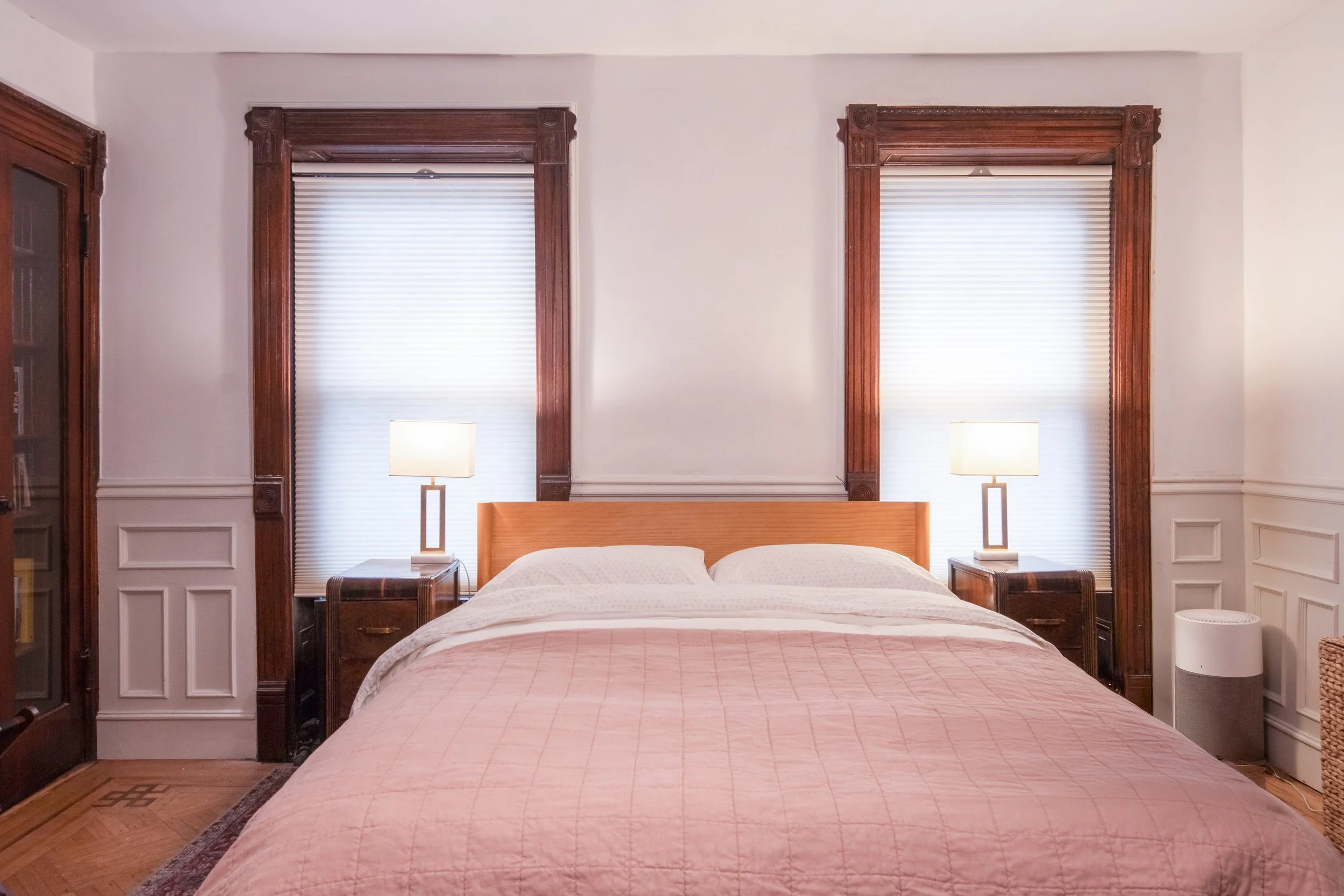 A cozy bedroom with a queen-size bed, pink quilt, and white pillows, flanked by two wooden nightstands with lamps, and two large windows with white blinds and ornate wooden frames.
