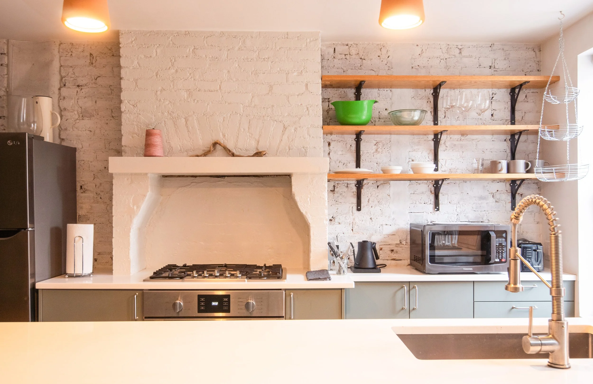 Modern kitchen with white brick walls, open wooden shelves holding bowls, glasses, and mugs, black and stainless steel appliances, and a brass faucet over a sink.