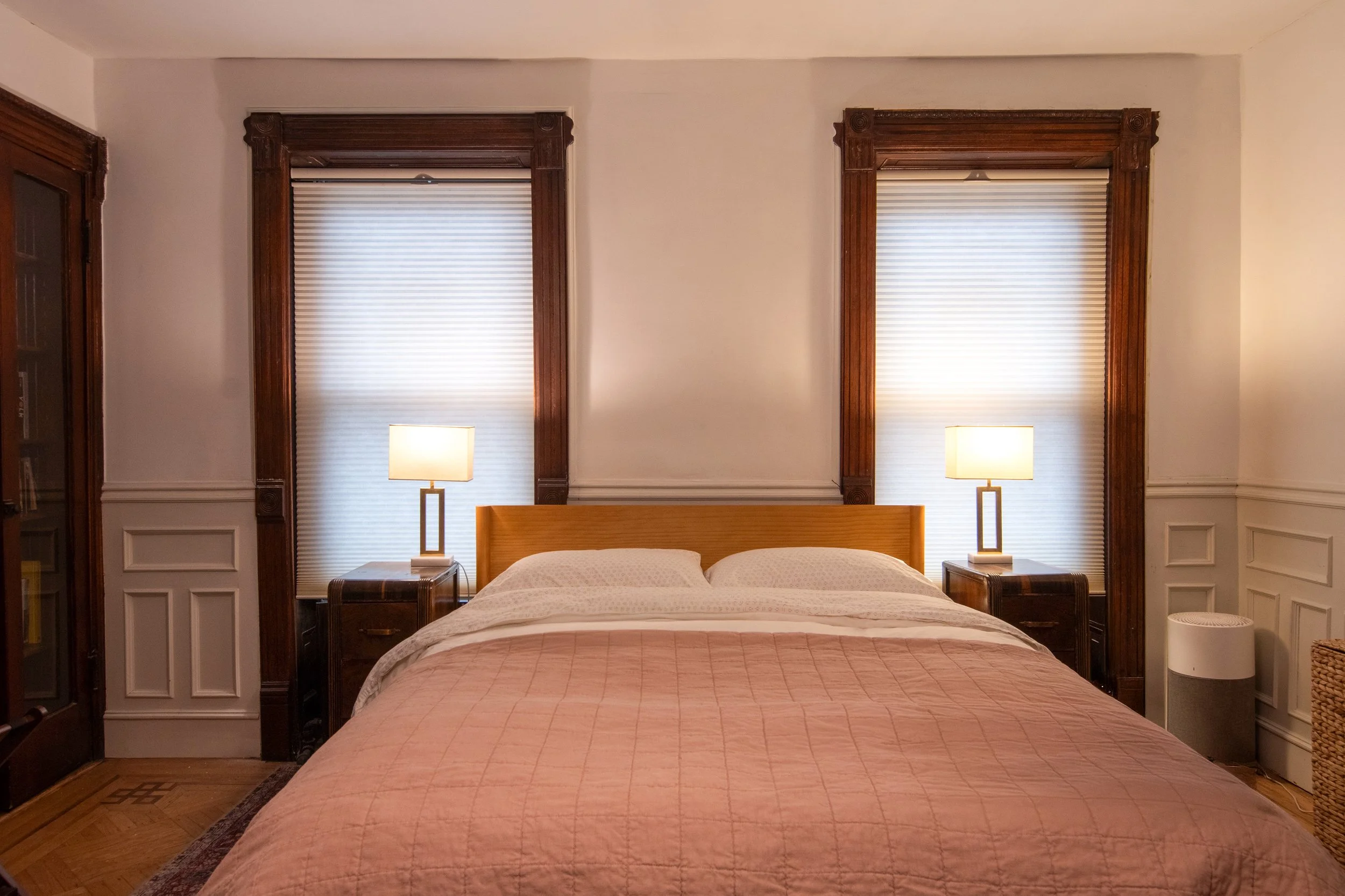 A cozy bedroom with a king-size bed, pink quilt, and white pillows, flanked by two wooden nightstands with lamps, and two large windows with white blinds and ornate wooden frames.