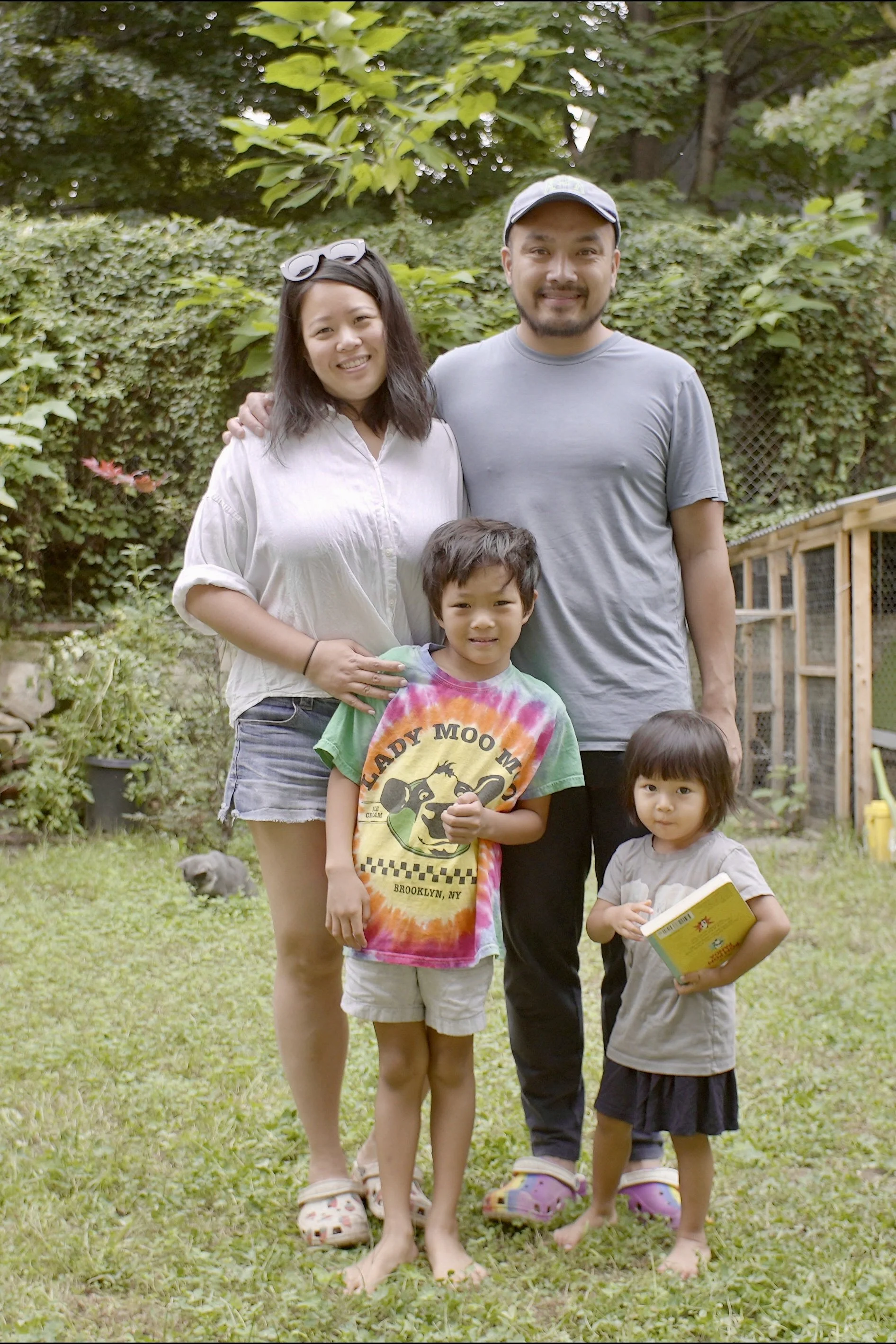 A family of five in a lush backyard, standing on grass with trees and a wooden fence in the background. The family includes a woman, a man, a boy, and a girl, with a gray cat in the background.