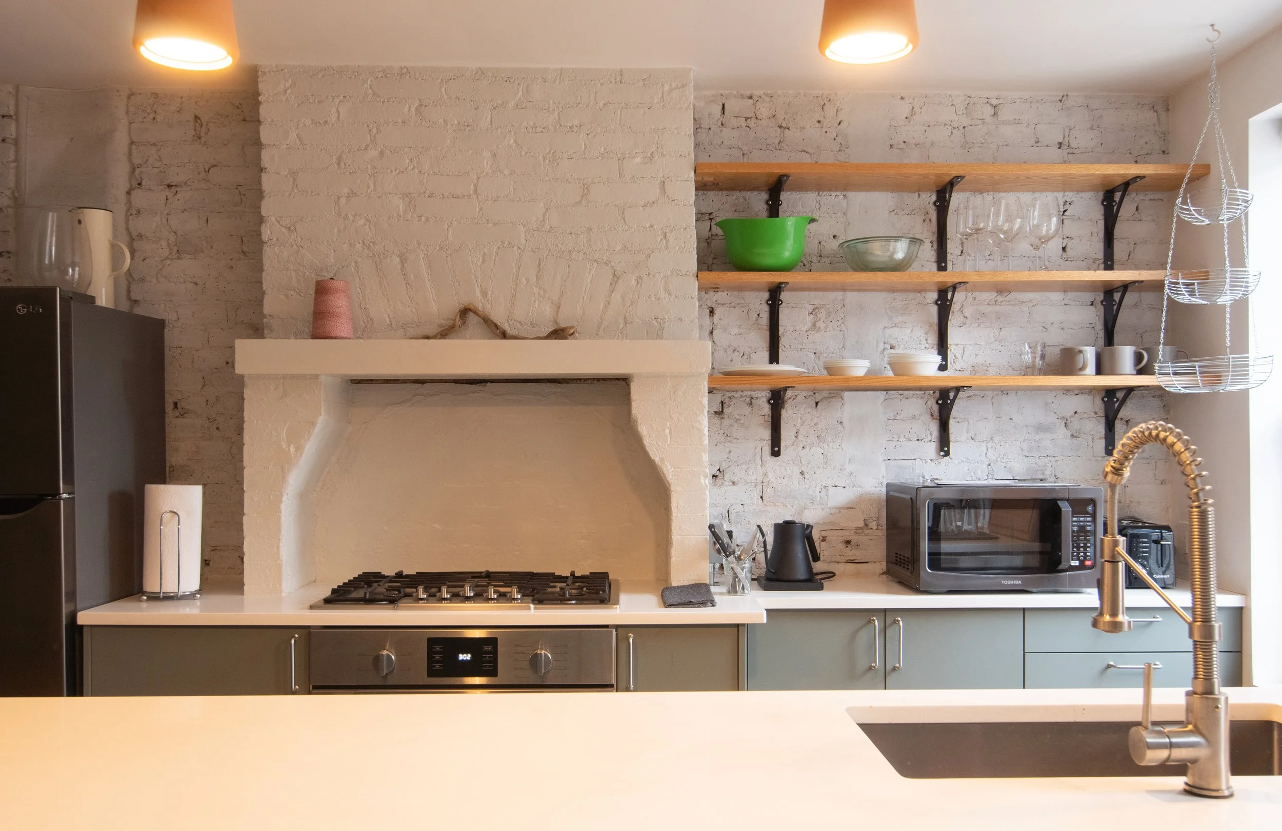 Modern kitchen with white brick walls, open wooden shelves holding bowls, glasses, and mugs, black and stainless steel appliances, and a brass faucet over a sink.