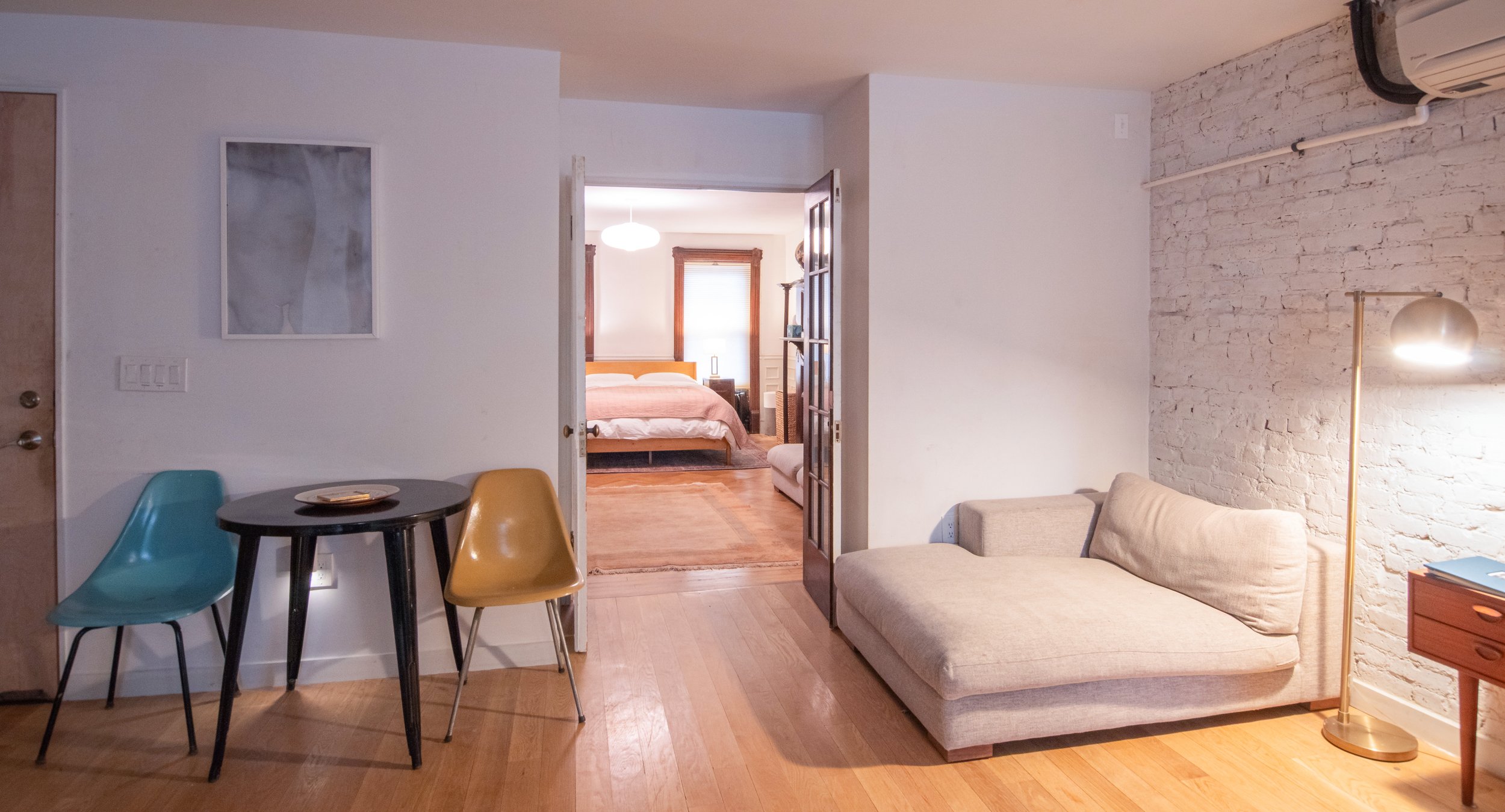 Living room with a beige sofa, floor lamp, small wooden table with blue and yellow chairs, and a framed picture on a white wall. Through an open door, a bedroom with a bed, window, and a pendant light is visible.