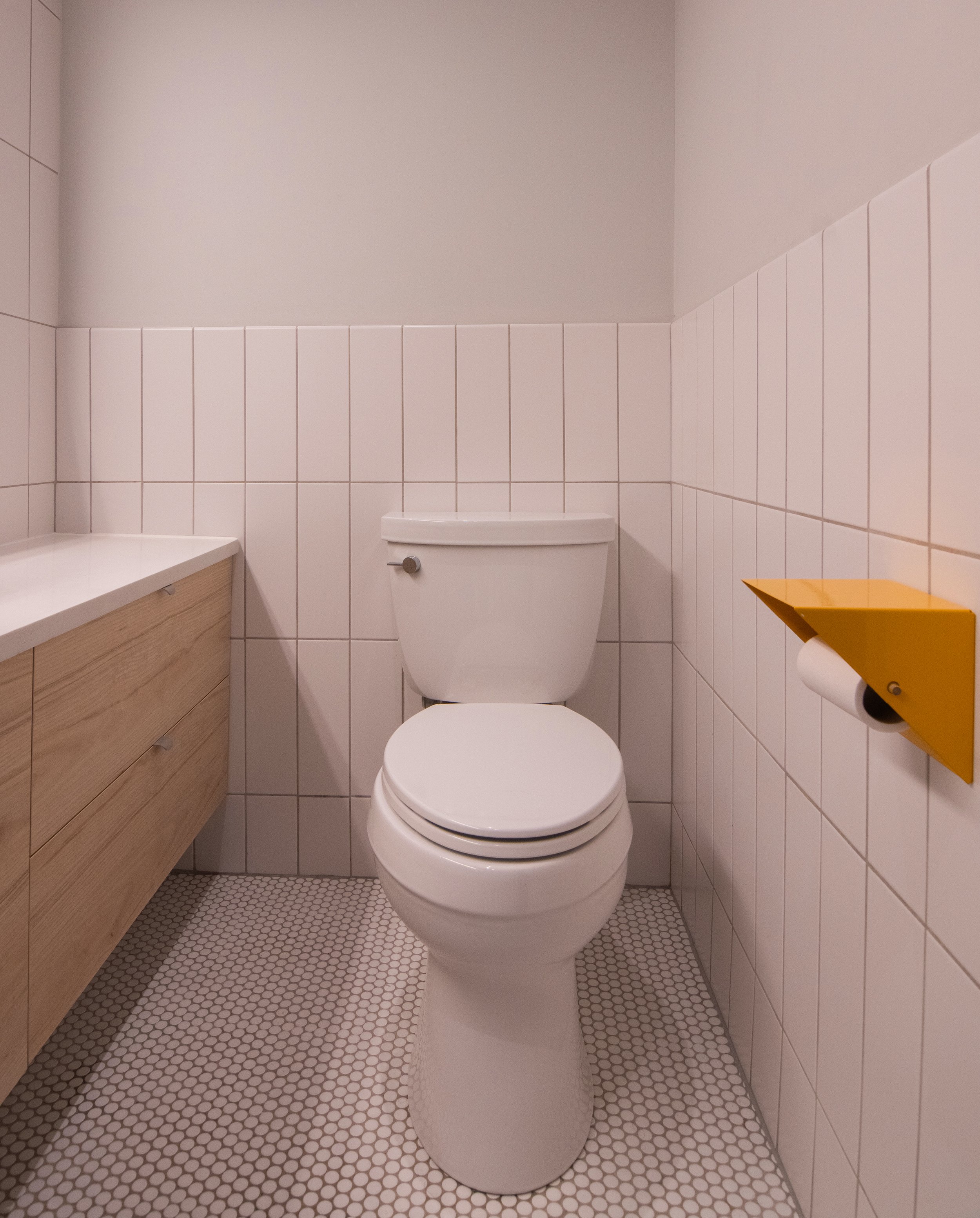 A small bathroom with a white toilet, light-colored wooden cabinet on the left, white tiled walls, and a yellow toilet paper holder on the right wall.